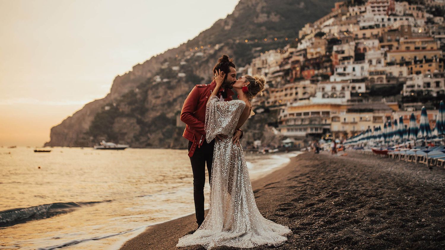 Couple in wedding attire sharing a romantic moment on a beach at sunset with Positano cliffside village backdrop.