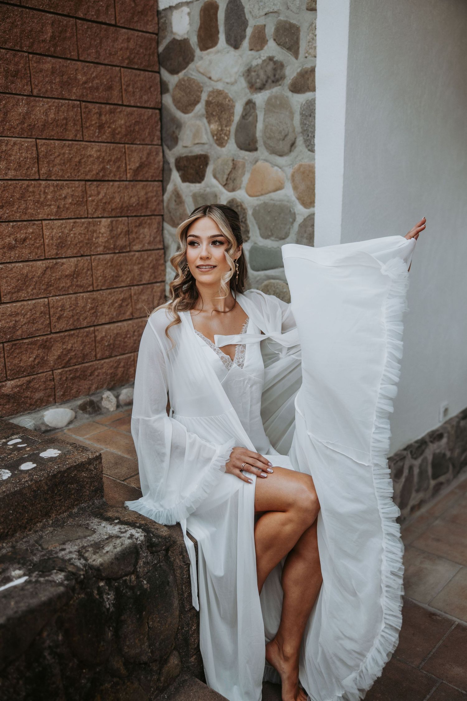Elegant bride poses in flowing white chiffon robe on stone steps, showcasing boho bridal fashion.