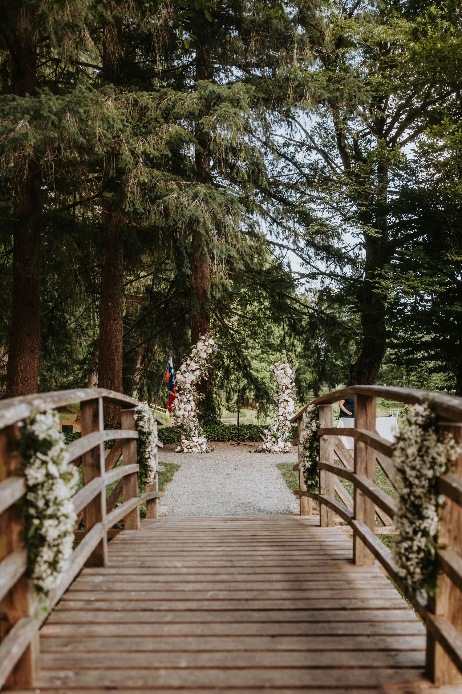 Wooden bridge adorned with white floral arrangements leads to a romantic garden wedding arch.