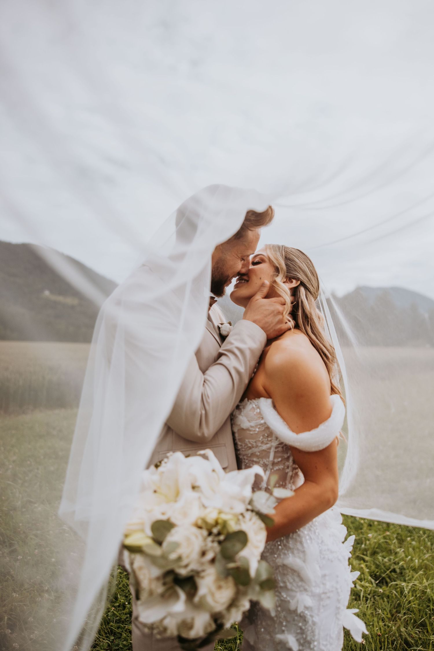 Bride and groom sharing a romantic kiss under a flowing white veil in a scenic outdoor wedding setting.
