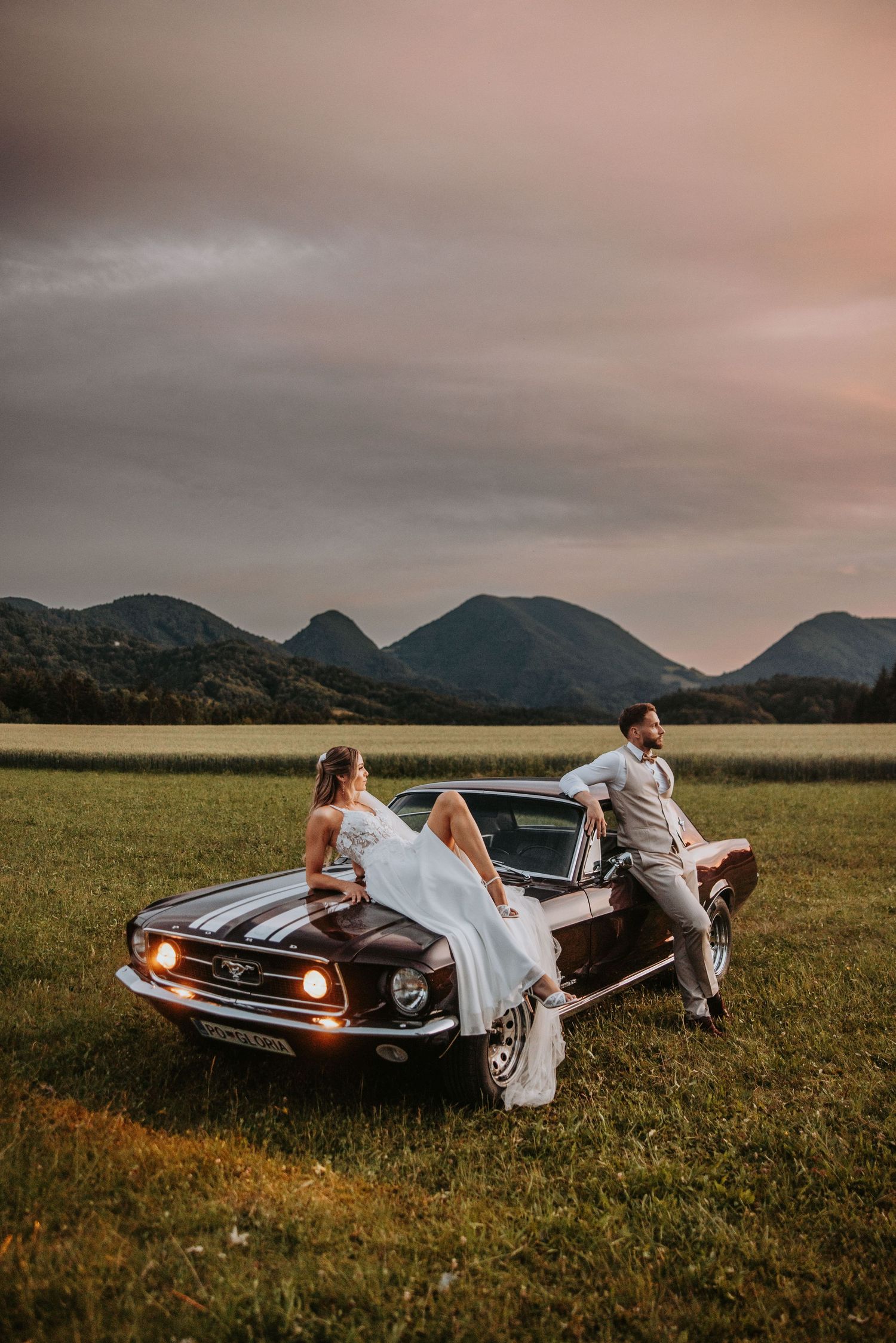 Bride and groom sit on vintage Ford Mustang hood in scenic mountain field at dusk.