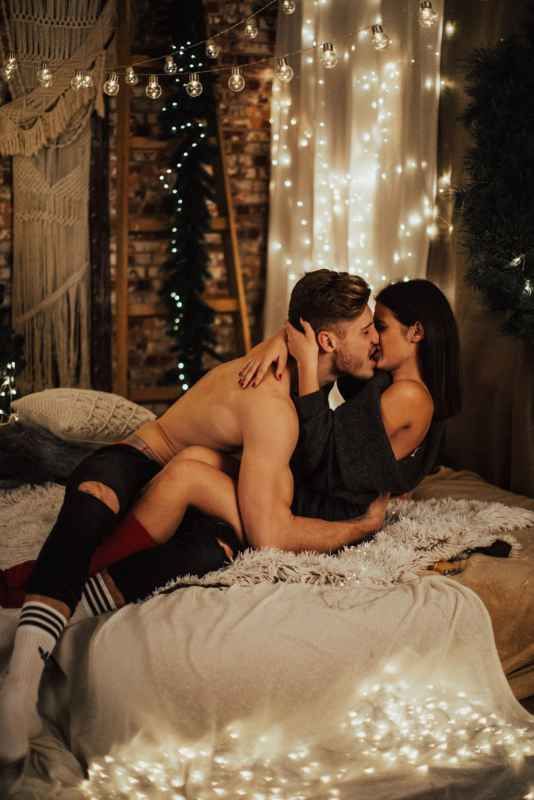 Romantic couple kissing on a bed surrounded by warm fairy lights in a cozy decorated room.