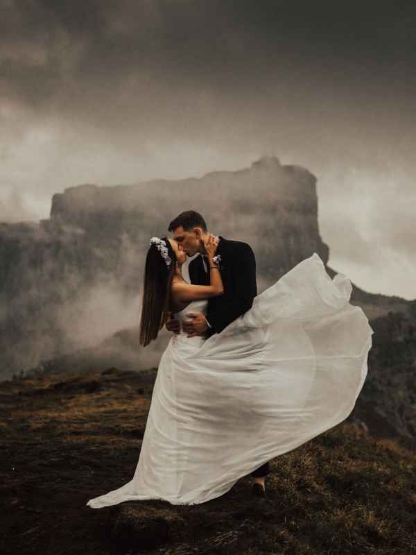 Couple kissing on misty mountain cliff, bride's white gown flowing dramatically in wind during romantic outdoor wedding.