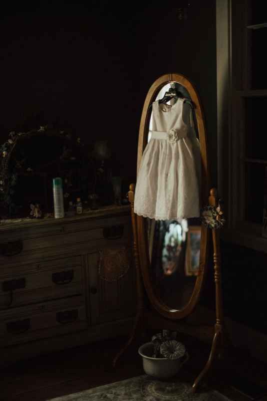 White flower girl dress hanging on wooden oval mirror in dimly lit vintage bedroom.