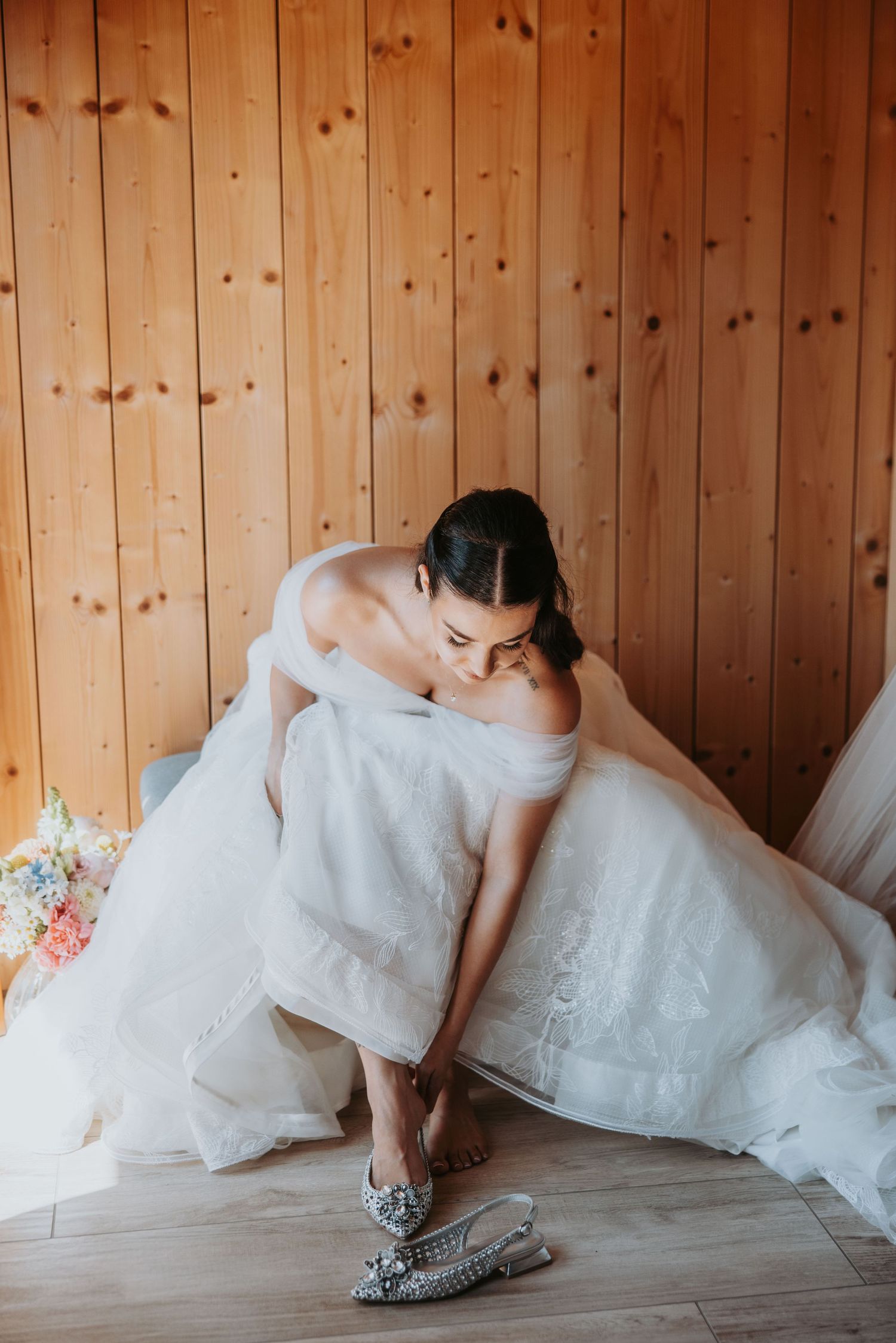 Bride in white off-shoulder gown putting on silver strappy heels on wooden floor against pine wall.