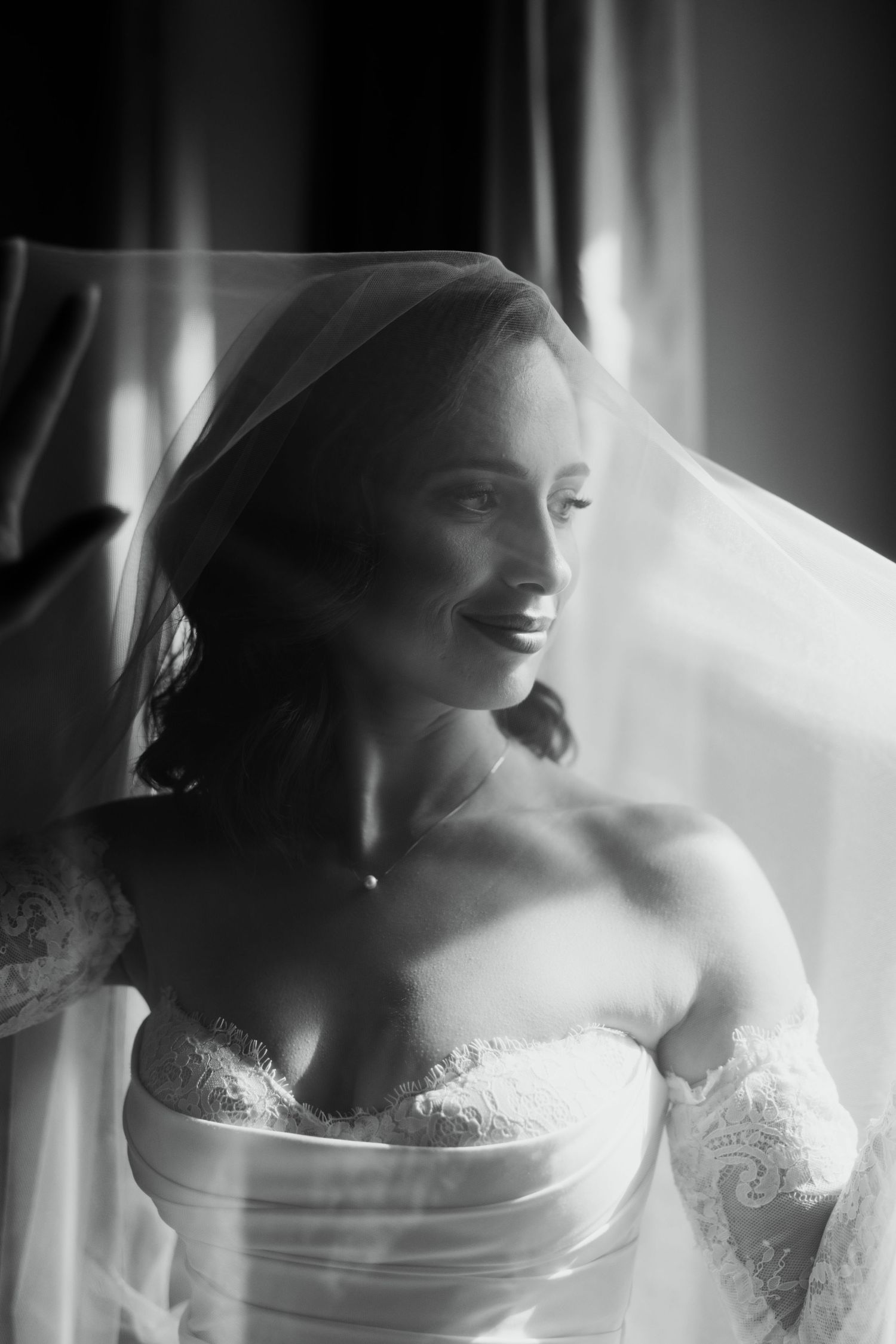 Beautiful bride in strapless gown and veil smiling by window, black and white.