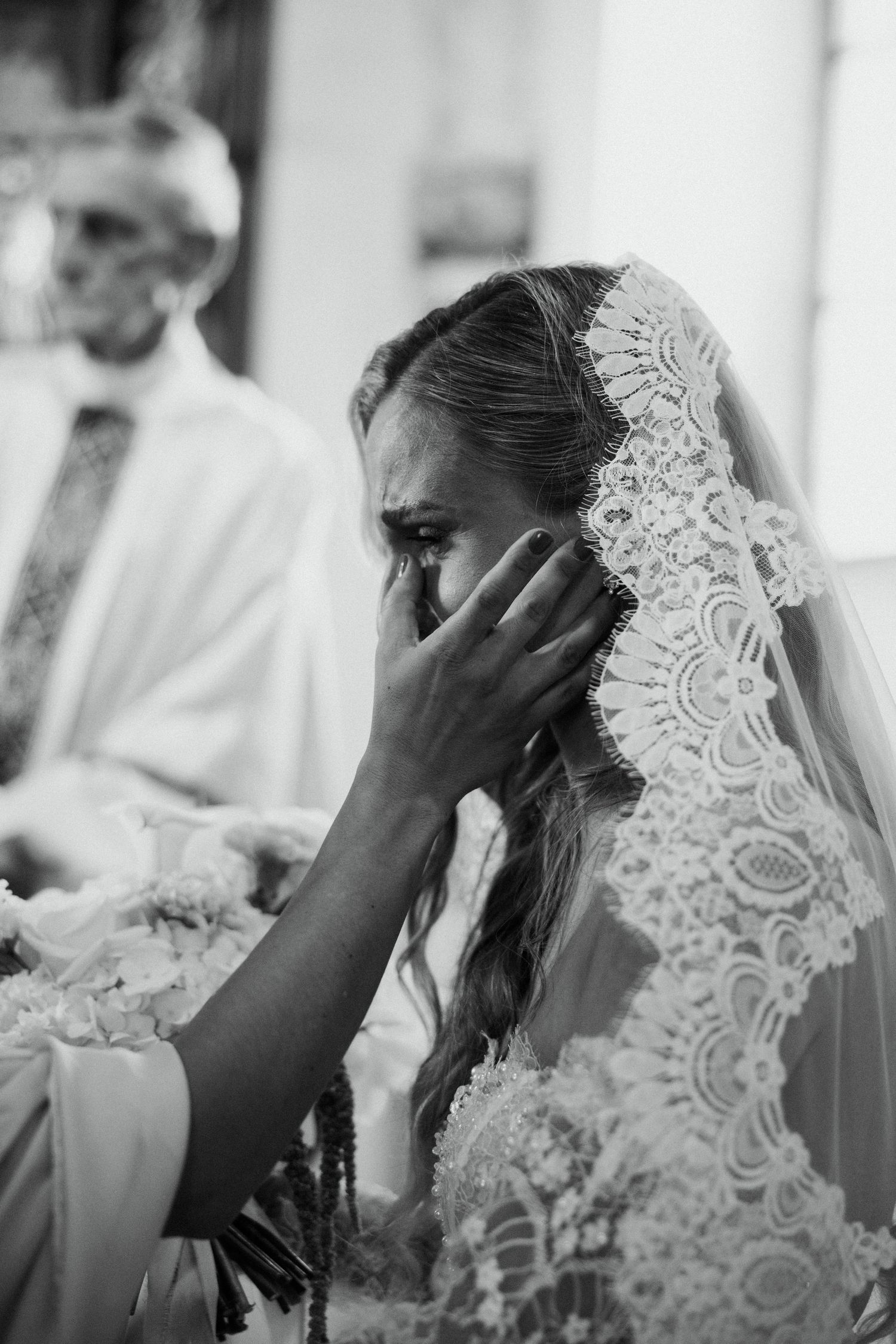 Emotional bride in lace veil wipes tears during church wedding ceremony in black and white photo.