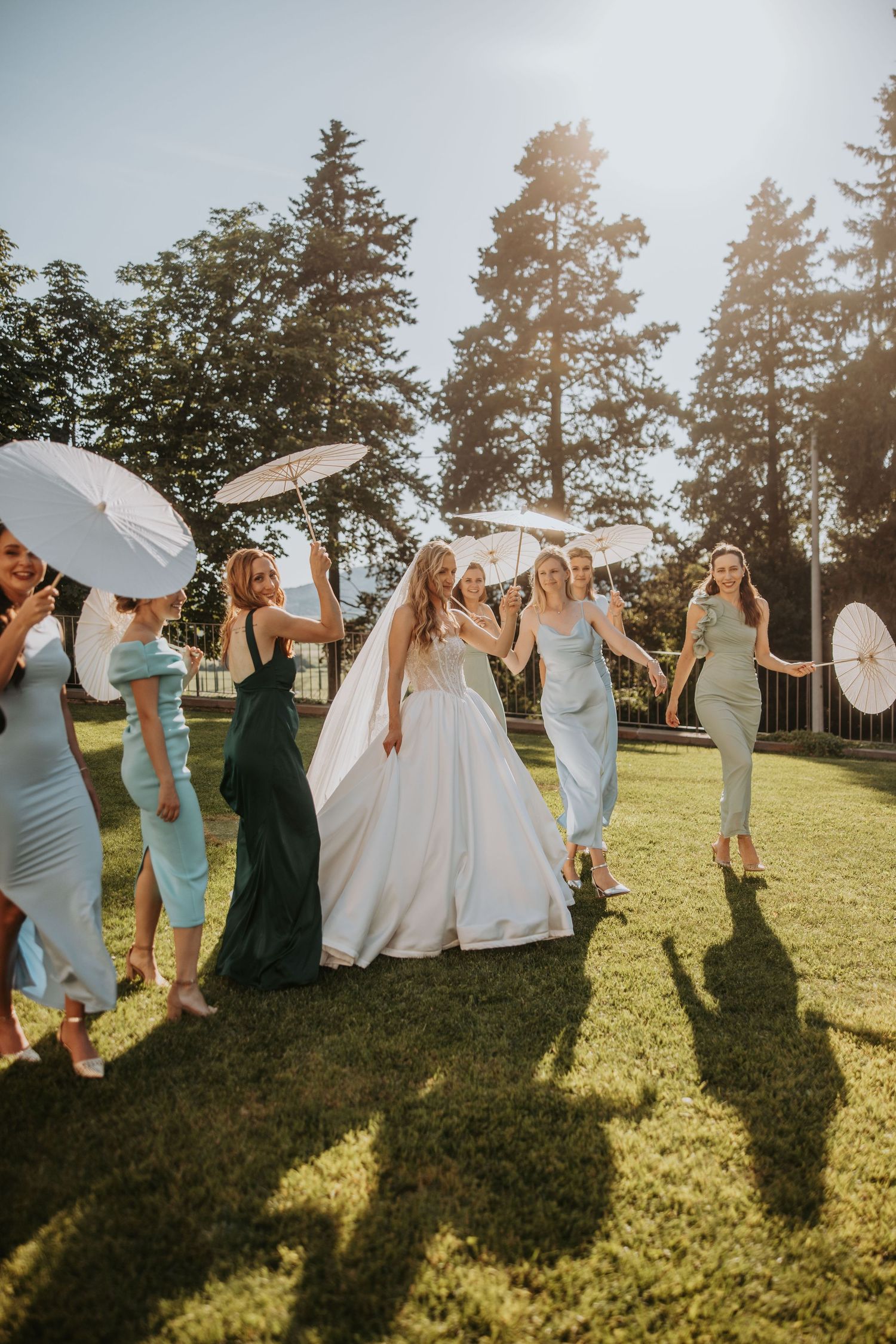 Bride and bridesmaids in blue dresses walking joyfully on green lawn with white parasols in sunlight.