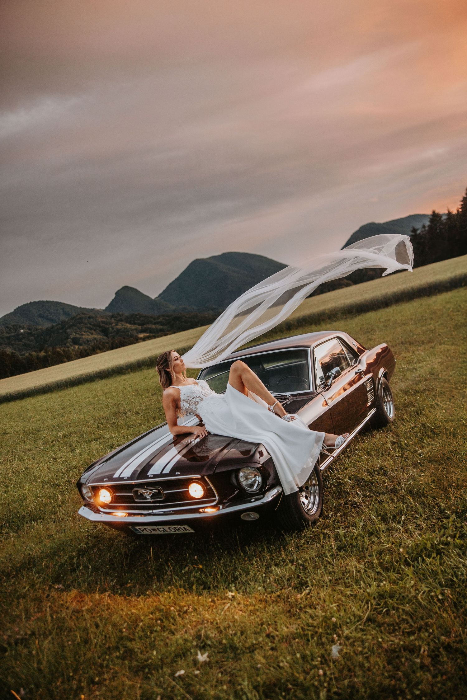 Bride in white dress posing on vintage black Ford Mustang hood at sunset with veil flowing in wind.