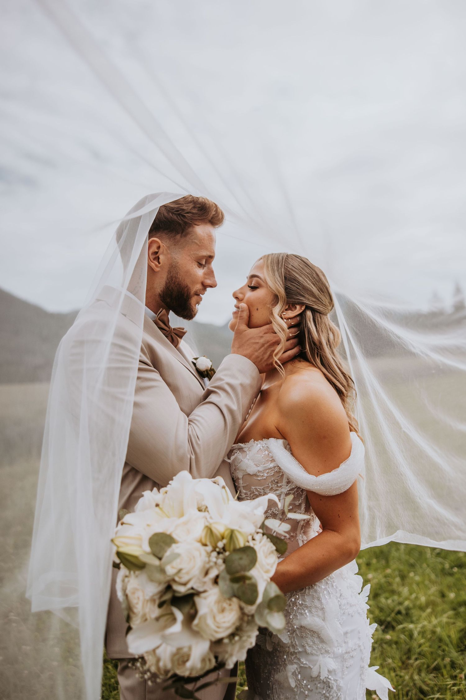 Romantic bride and groom sharing an intimate moment under flowing veil in scenic field.