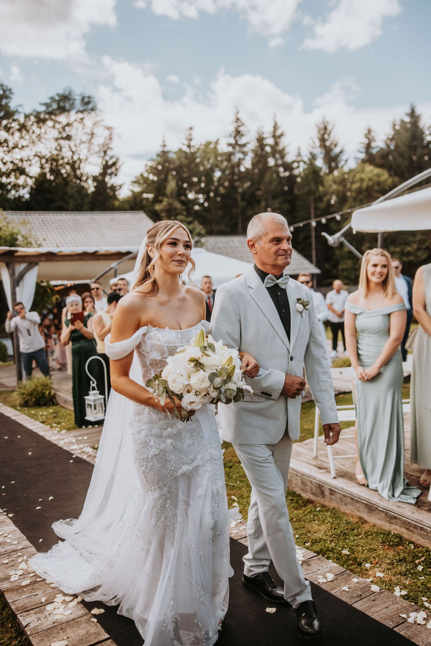 Bride in lace gown walks down outdoor aisle with escort holding white bouquet at garden ceremony.
