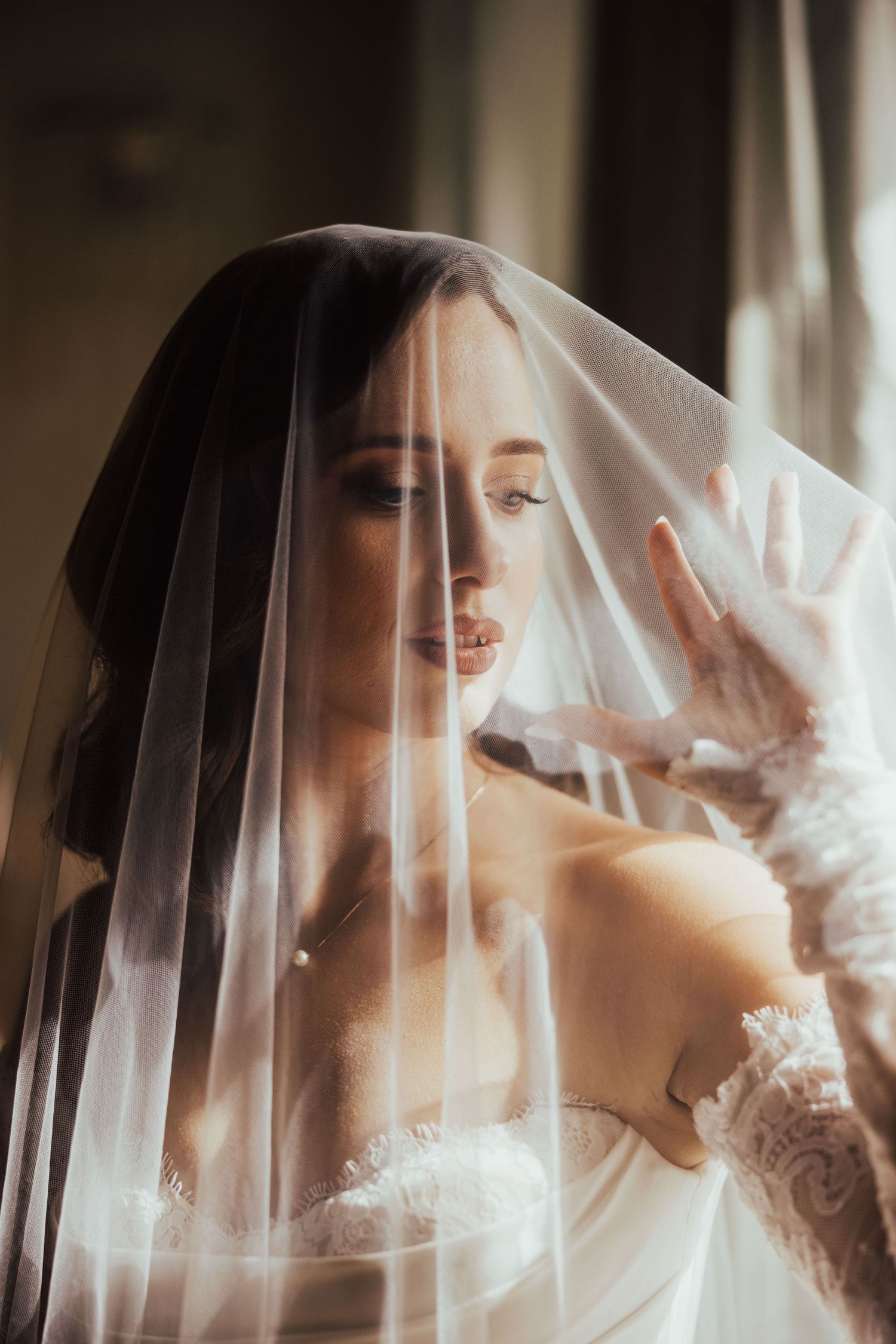 Beautiful bride in lace wedding dress lifting her veil near a sunlit window on her wedding day.