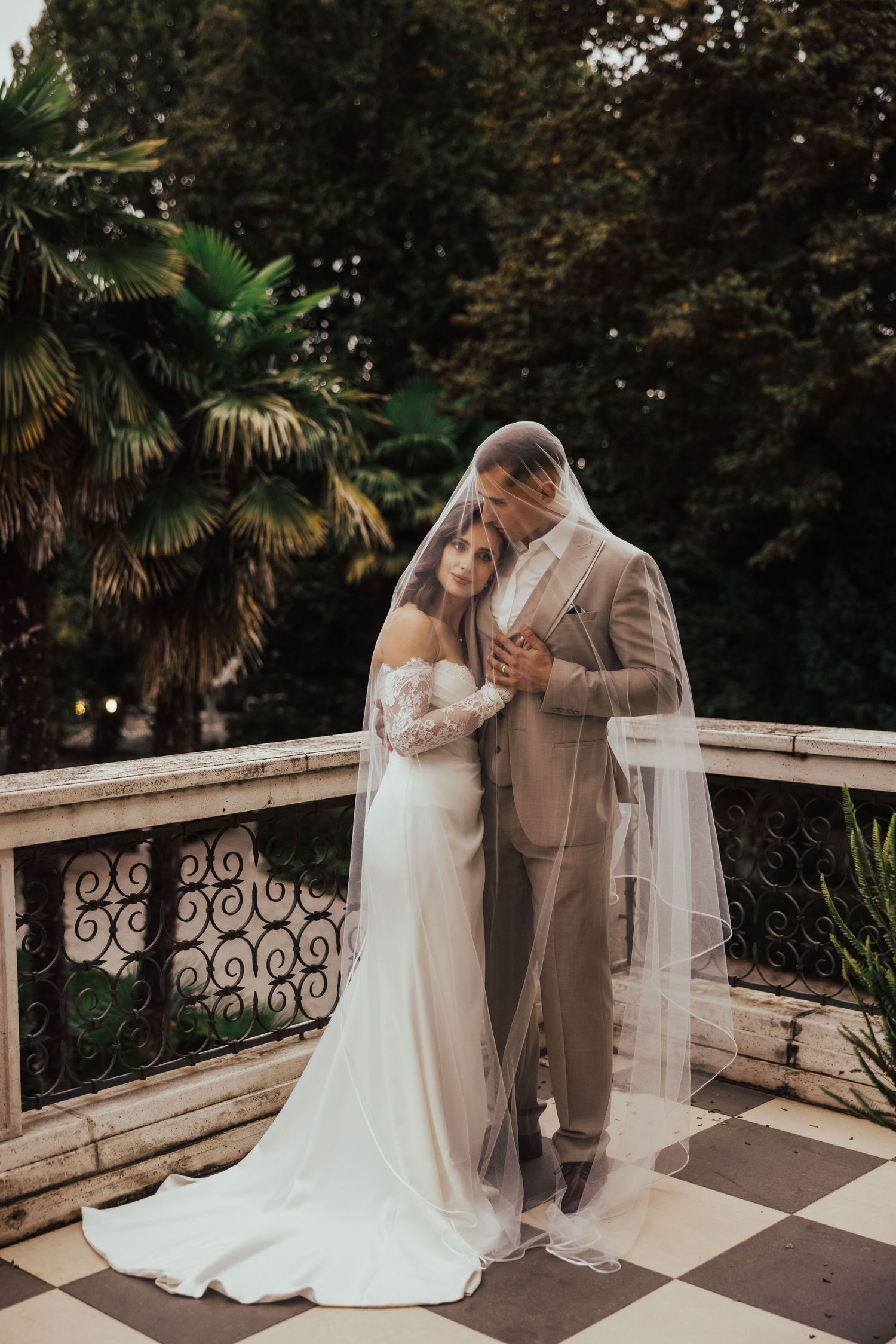 Bride and groom sharing an intimate moment under a veil on an ornate balcony surrounded by trees.
