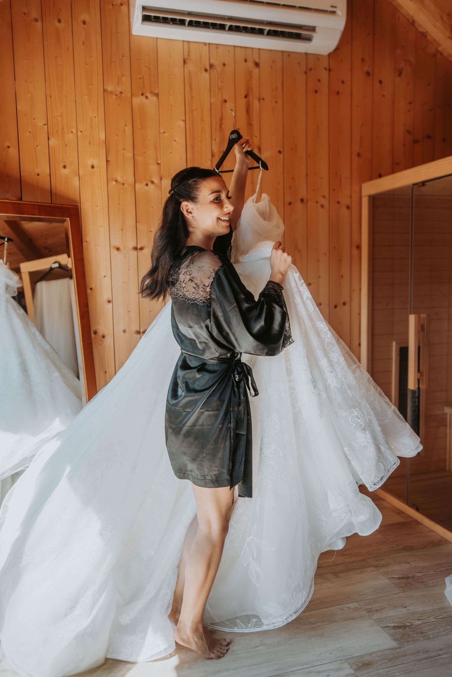 Bride in gray robe holding white wedding dress on hanger in wooden cabin bridal suite.