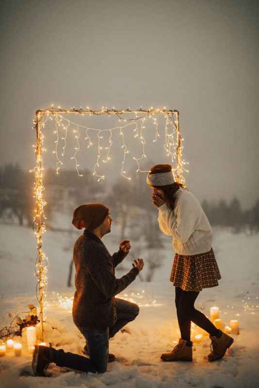 Romantic winter proposal under fairy light arch with candles in snow at dusk.