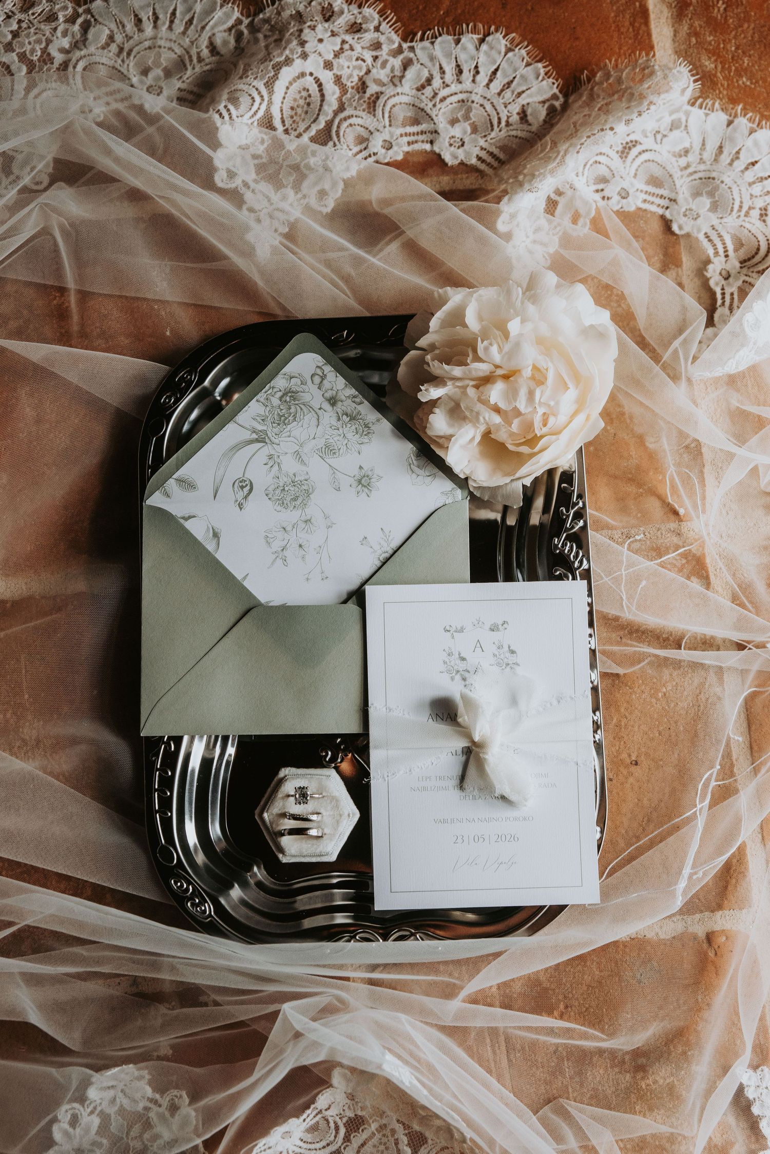 Elegant wedding flat lay with sage green envelope, floral stationery, ring box, and white peony on silver tray with lace veil.