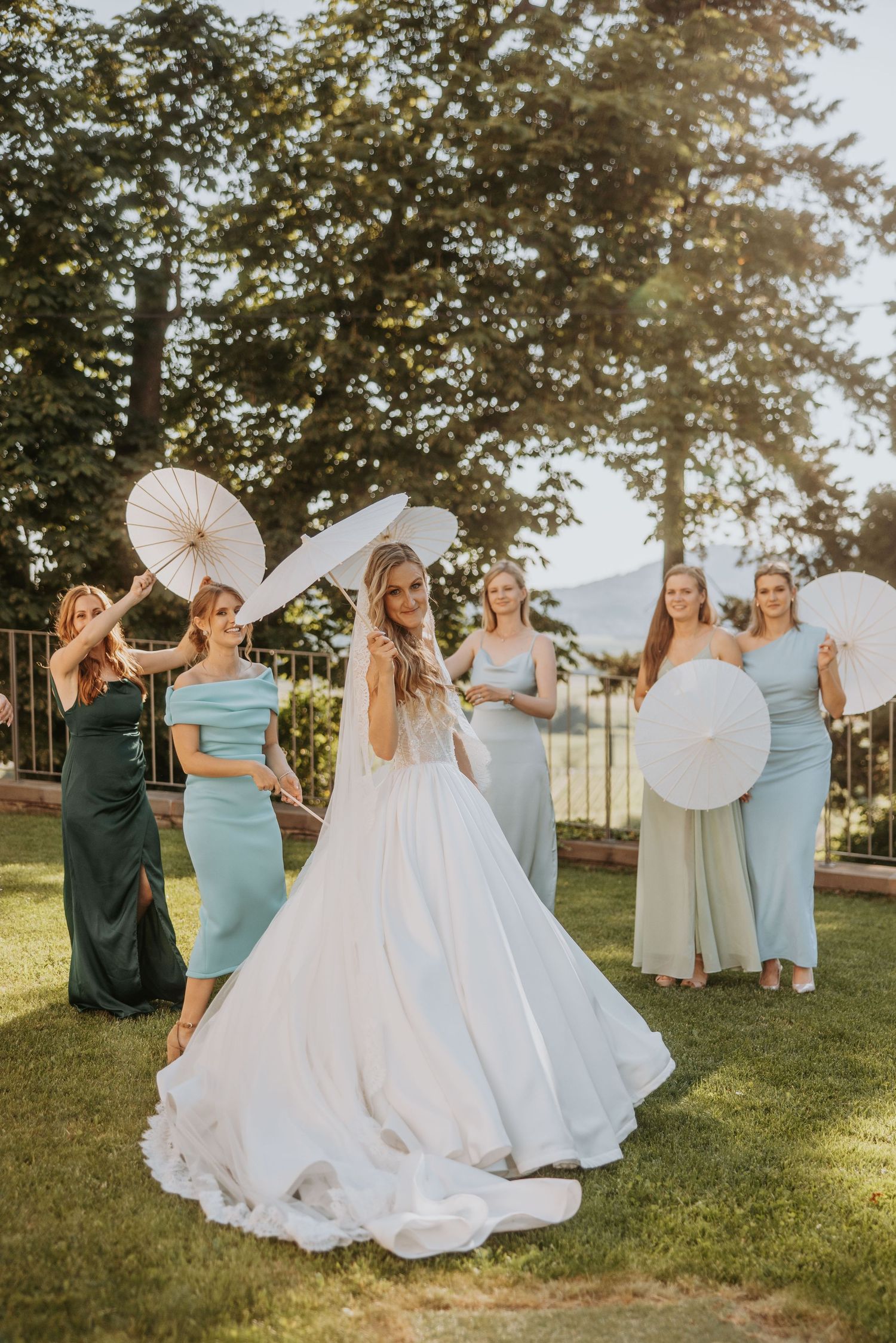 Bride twirls in white wedding gown while bridesmaids in sage green dresses hold white parasols outdoors.