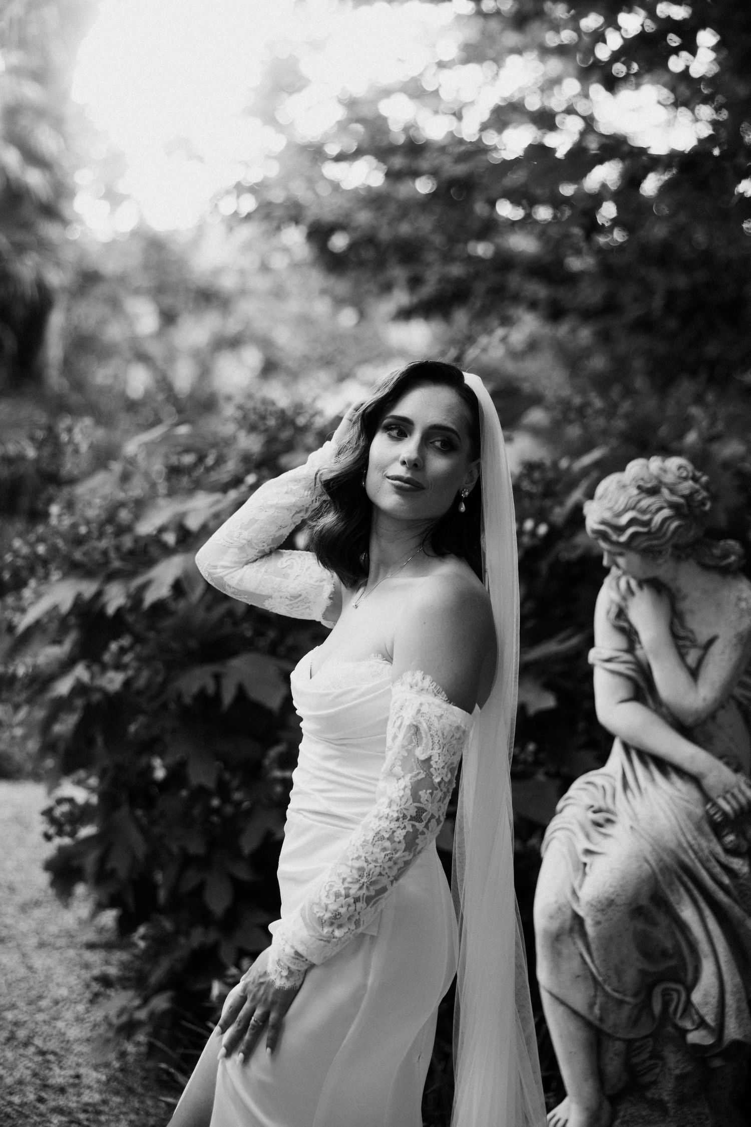Elegant bride in white gown with lace sleeves posing by garden statue.