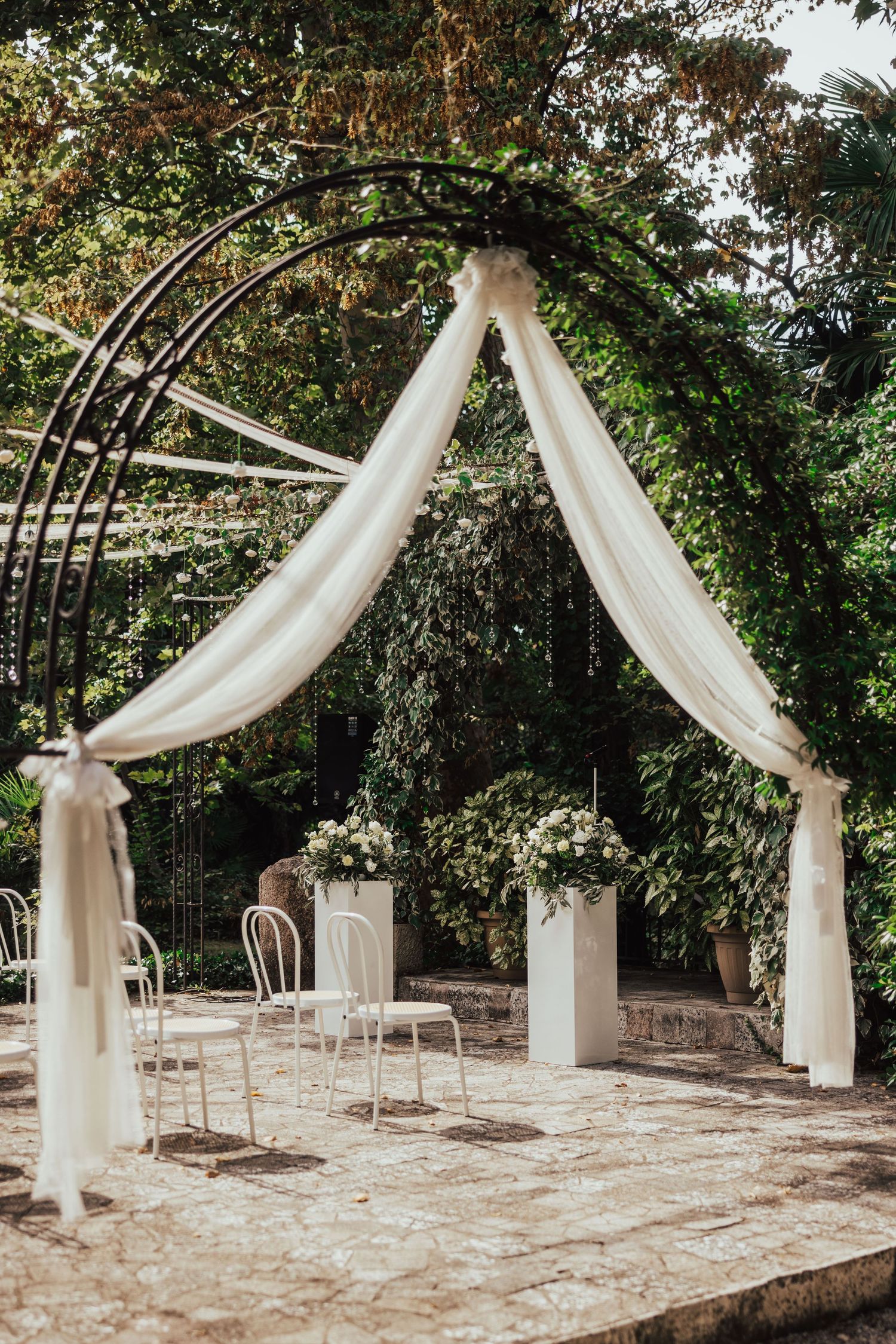 Outdoor wedding ceremony arch with white draping and floral arrangements in lush garden.