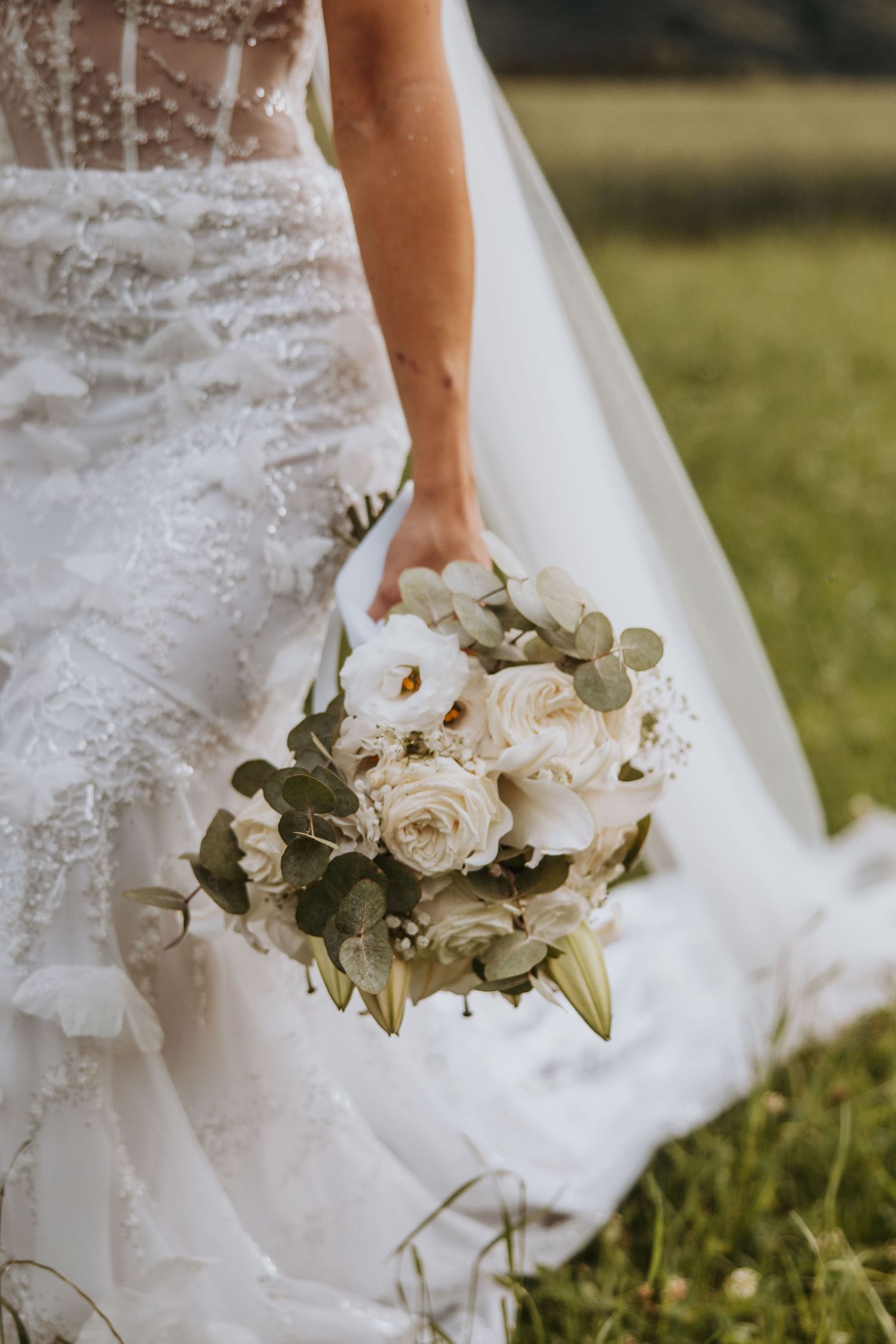 Bride holding a white and green bridal bouquet of roses and eucalyptus against lace wedding gown.