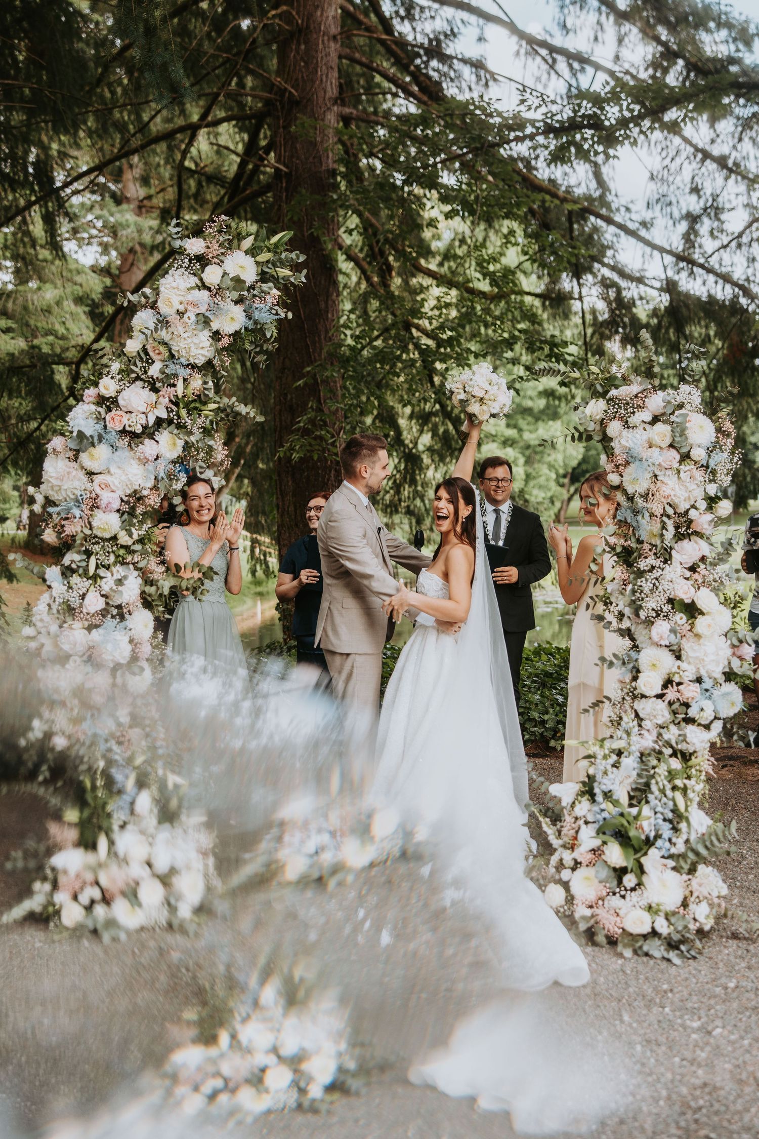 Joyful bride and groom celebrate under floral arch surrounded by wedding party outdoors.