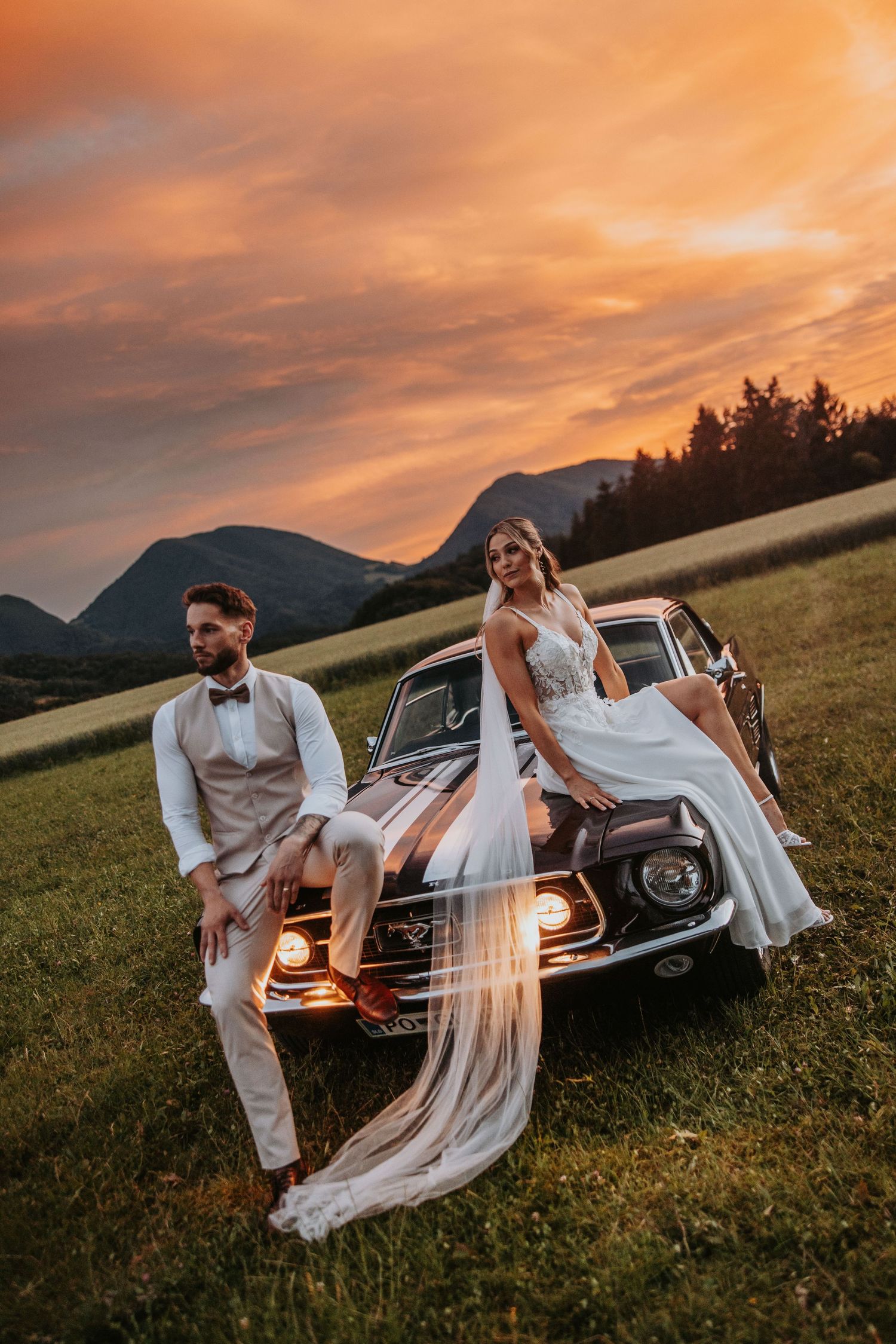 Bride and groom posing against vintage car at sunset with dramatic orange sky and mountains.