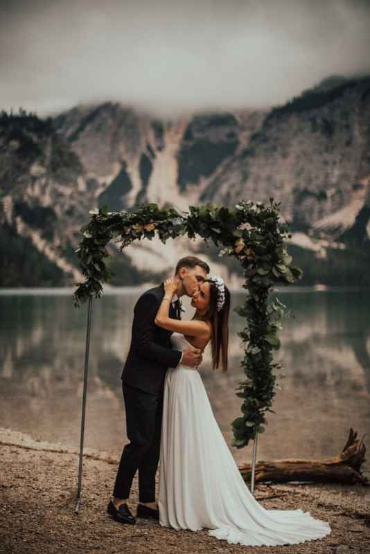 Couple kissing under floral arch at lakeside wedding with dramatic mountain scenery behind them.