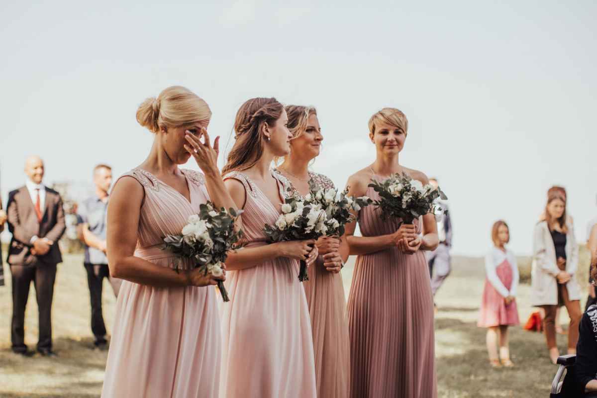 Bridesmaids in blush pink dresses holding white bouquets at an outdoor wedding ceremony.