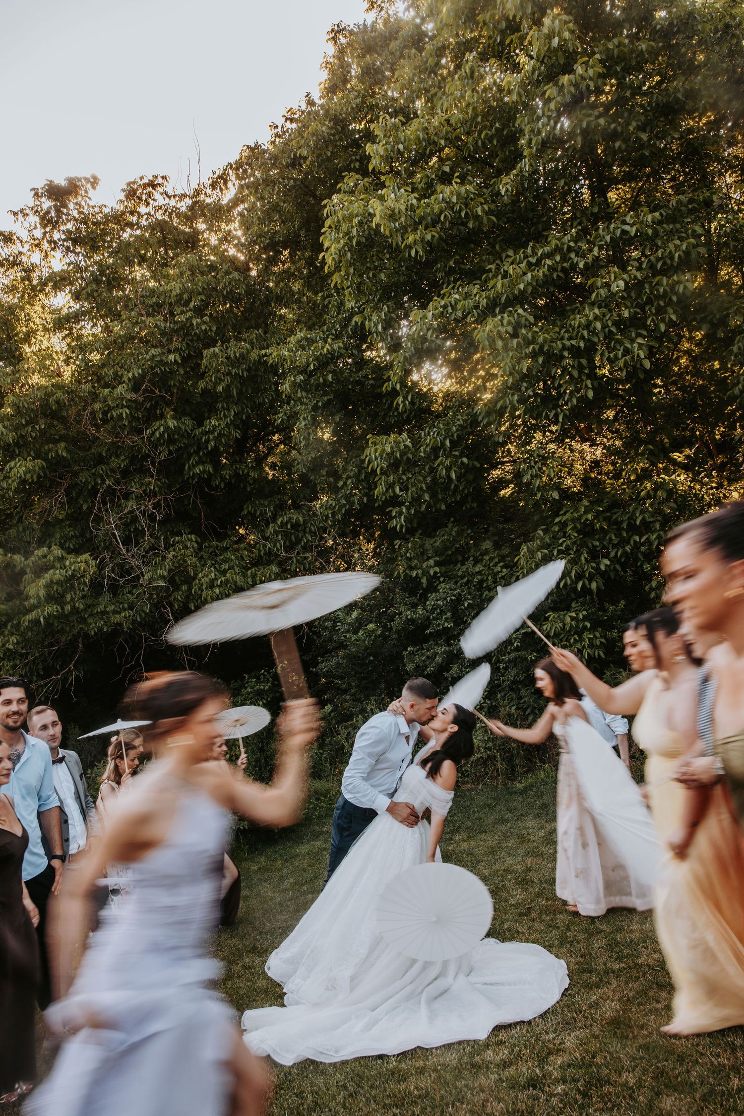 Bride and groom kissing surrounded by guests with parasols in outdoor wedding celebration.