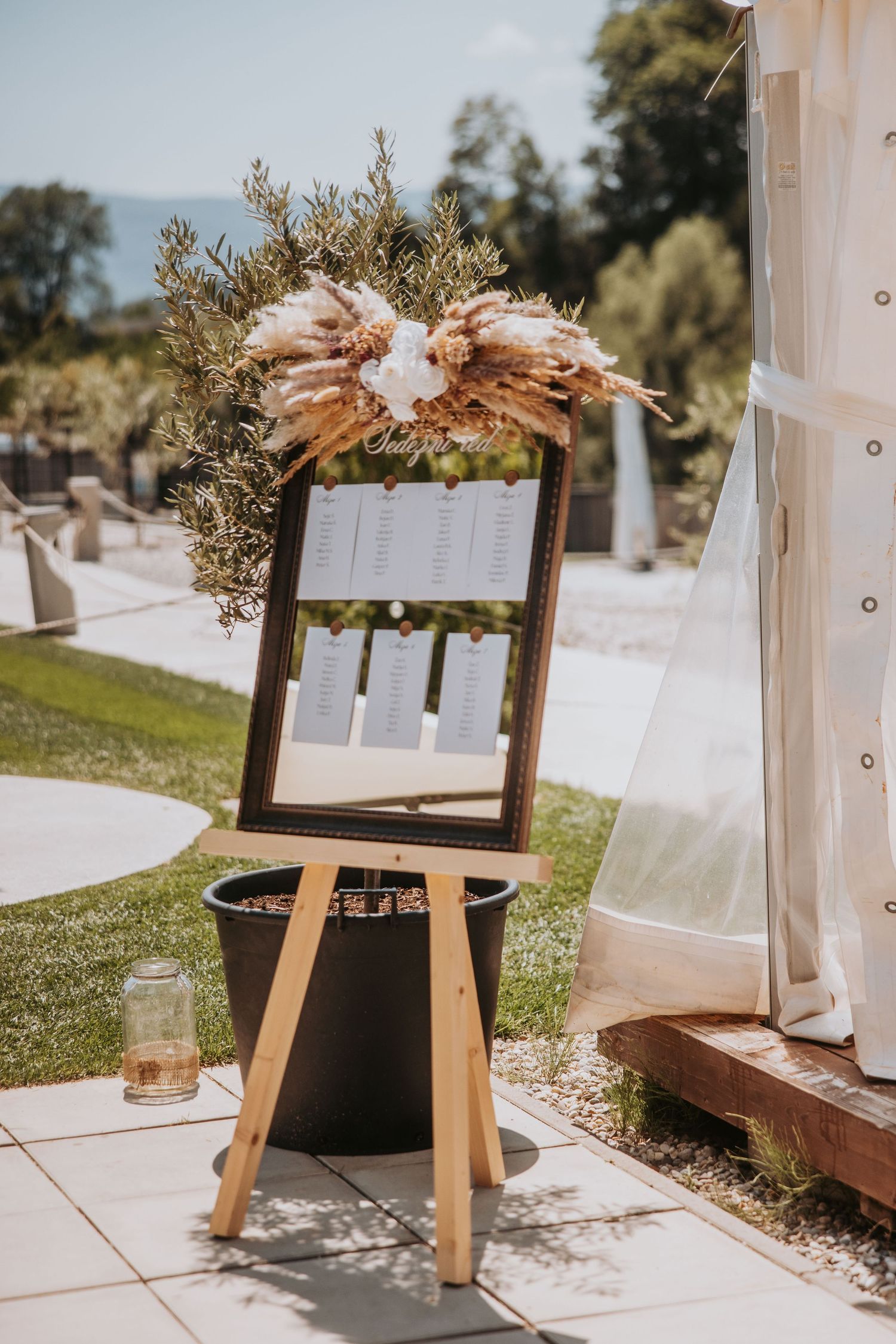 Outdoor wedding seating chart on wooden easel decorated with pampas grass and dried florals at sunny venue.