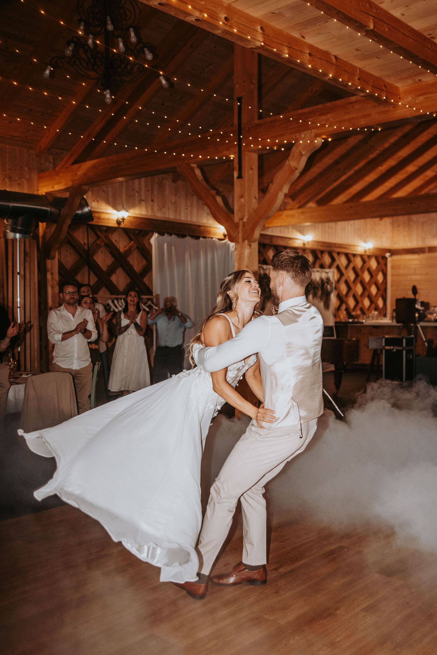Bride and groom sharing joyful first dance in rustic wooden barn with string lights and fog machine.