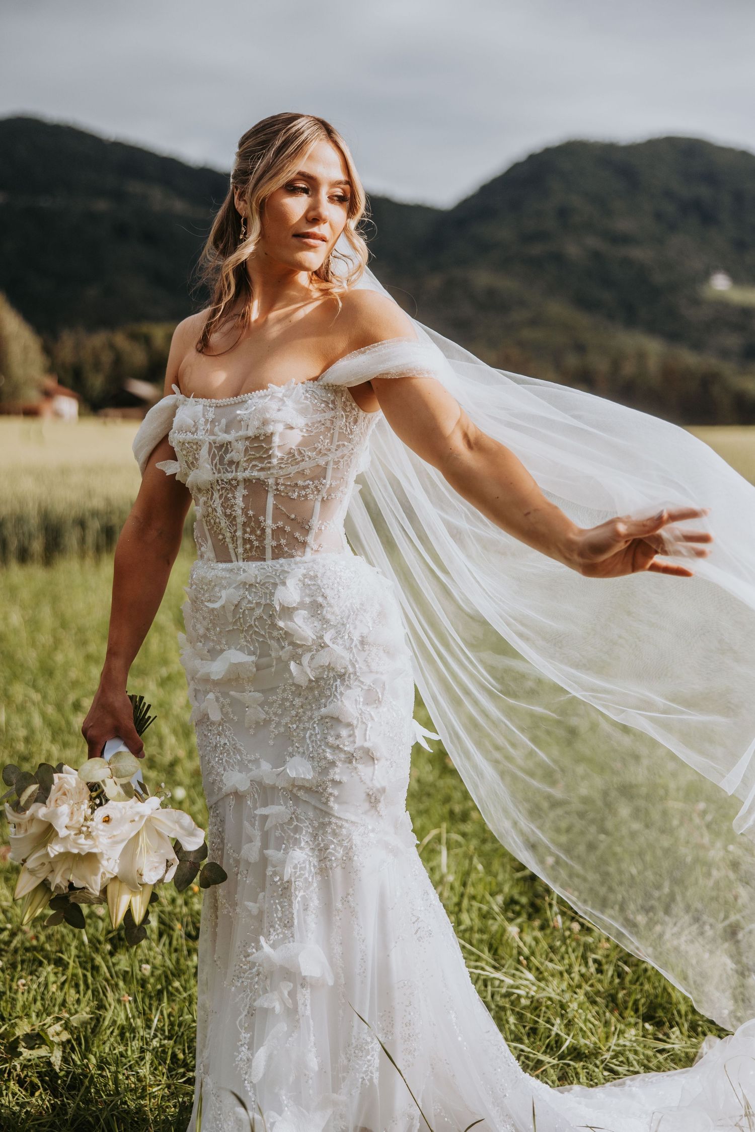 Bride in off-shoulder lace gown lets veil flow in mountain meadow while holding white floral bouquet.