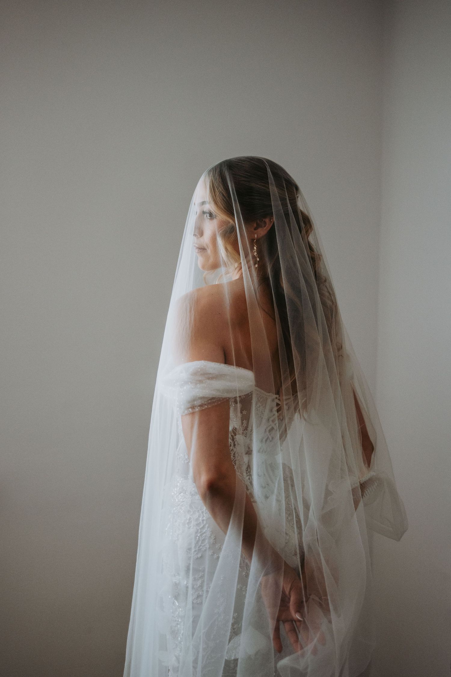 Elegant bride in a lace off-shoulder wedding dress draped under a long white veil, side profile view.