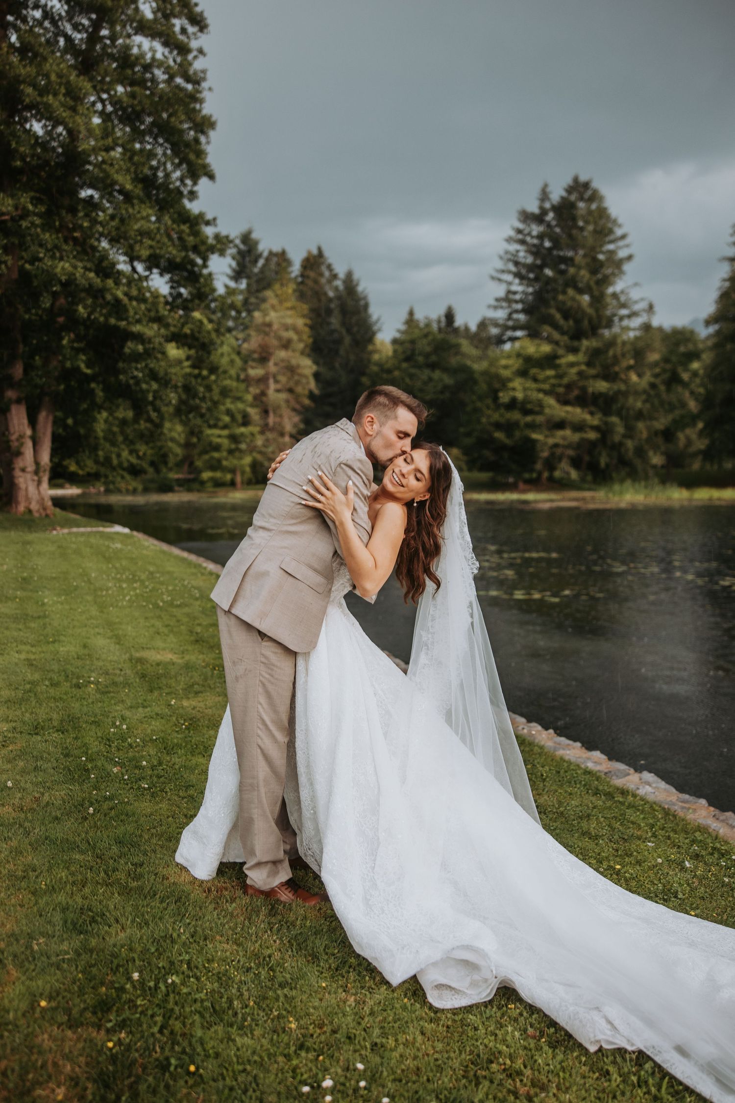 Bride and groom embracing by a serene lake surrounded by lush trees on their wedding day.