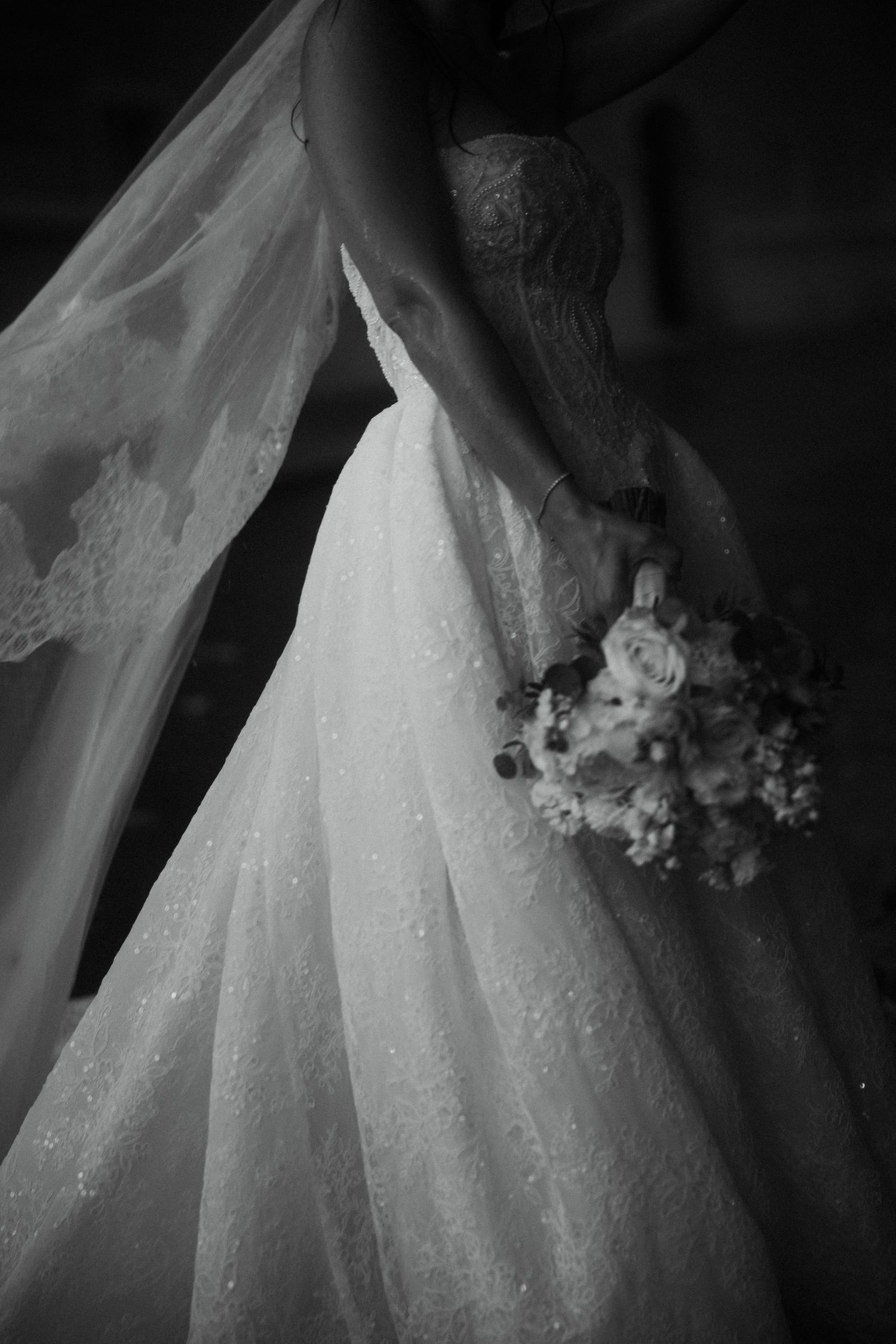 Black and white close-up of a bride in a lace wedding gown holding a delicate floral bouquet with a flowing veil.