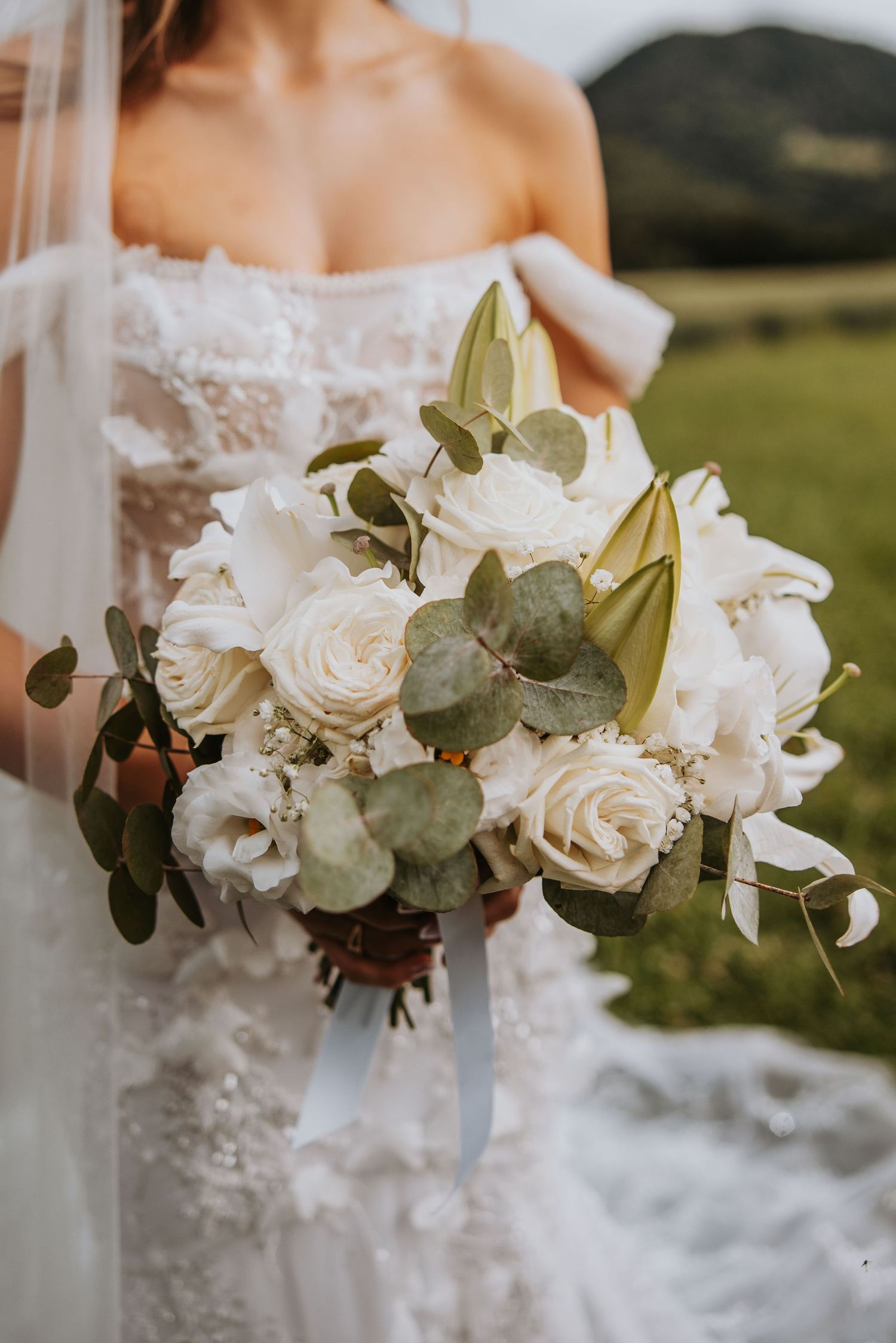 Bride holding a lush white rose and eucalyptus wedding bouquet with ribbon, standing in a green field.