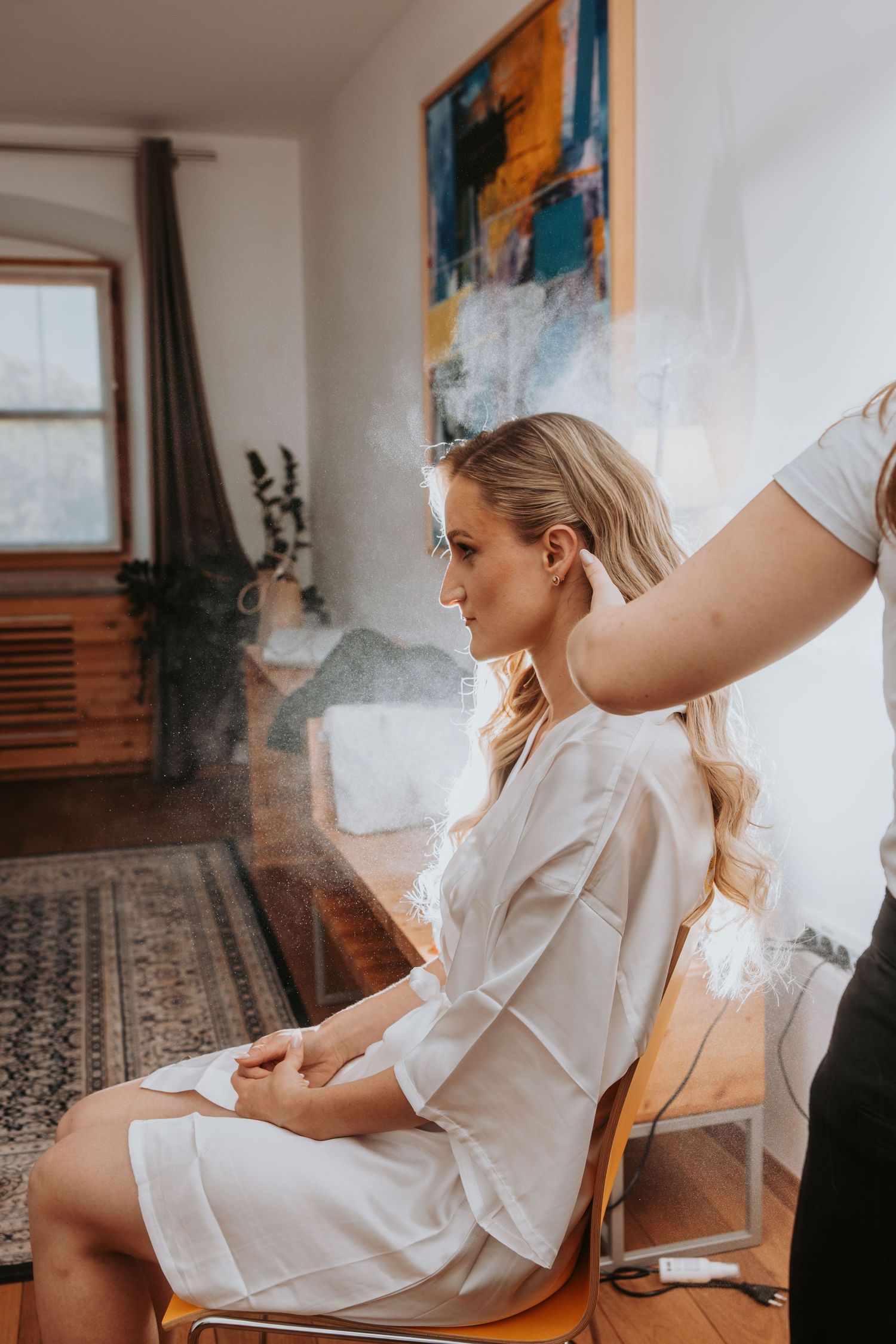 Bride in white robe getting hair styled with hairspray before her wedding.