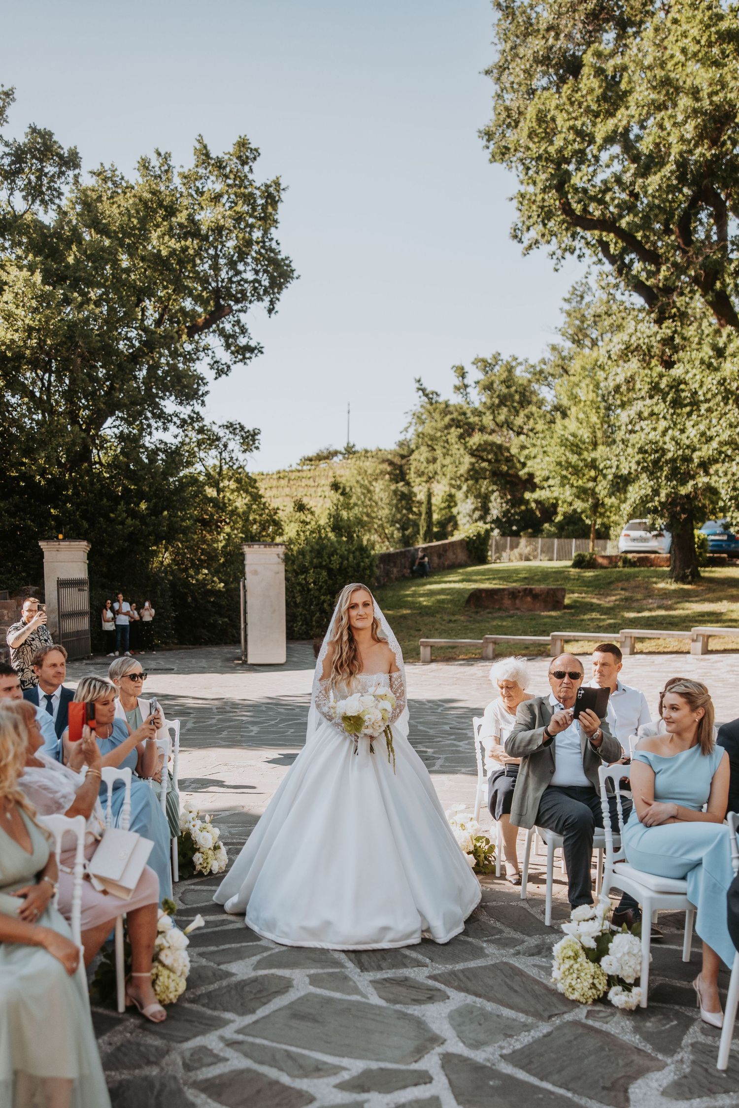 Bride in ballgown walking down outdoor aisle holding white bouquet with seated wedding guests on sunny day.