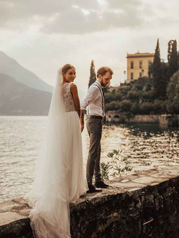 Bride and groom standing on stone wall overlooking lake with mountains and villa backdrop.