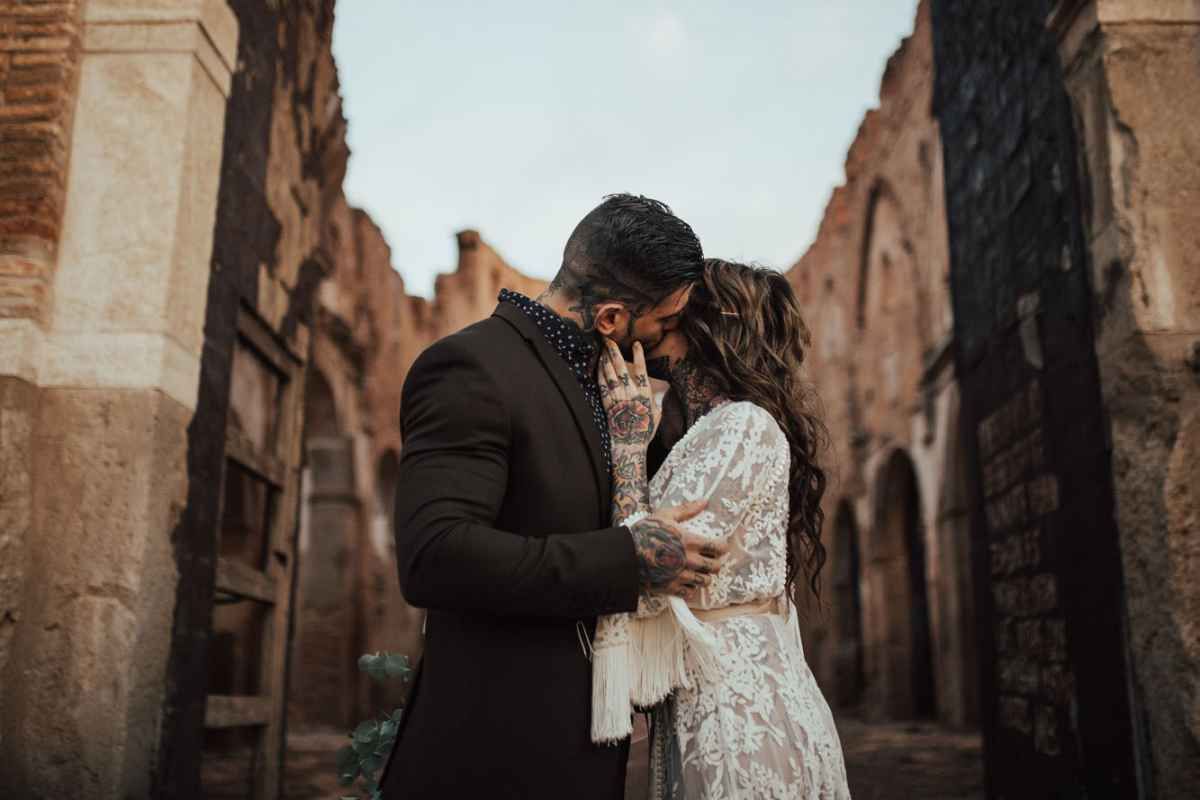 Couple sharing a romantic kiss in wedding attire among ancient stone ruins at golden hour.