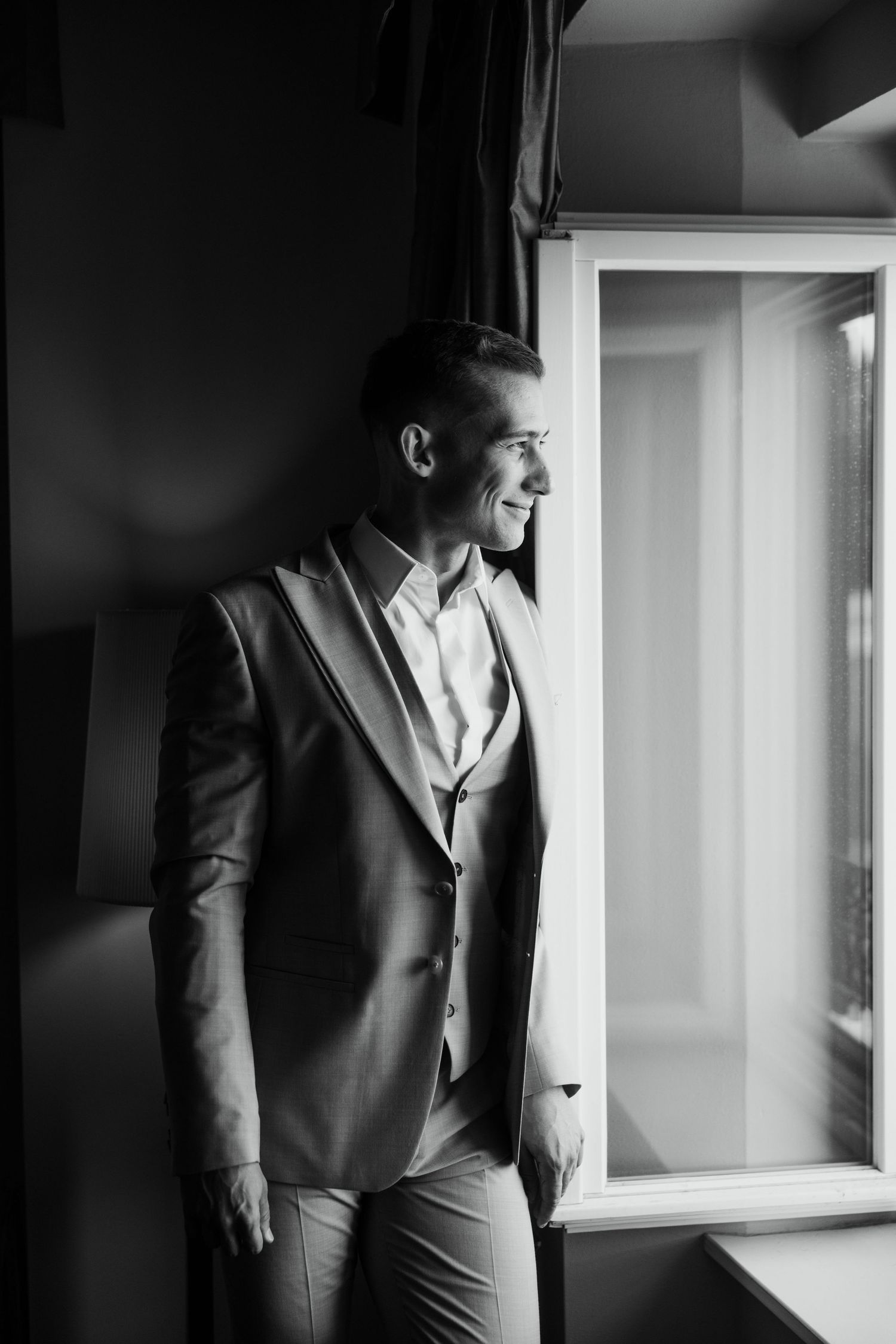 Elegant groom in beige suit smiling by window in black and white portrait photo.