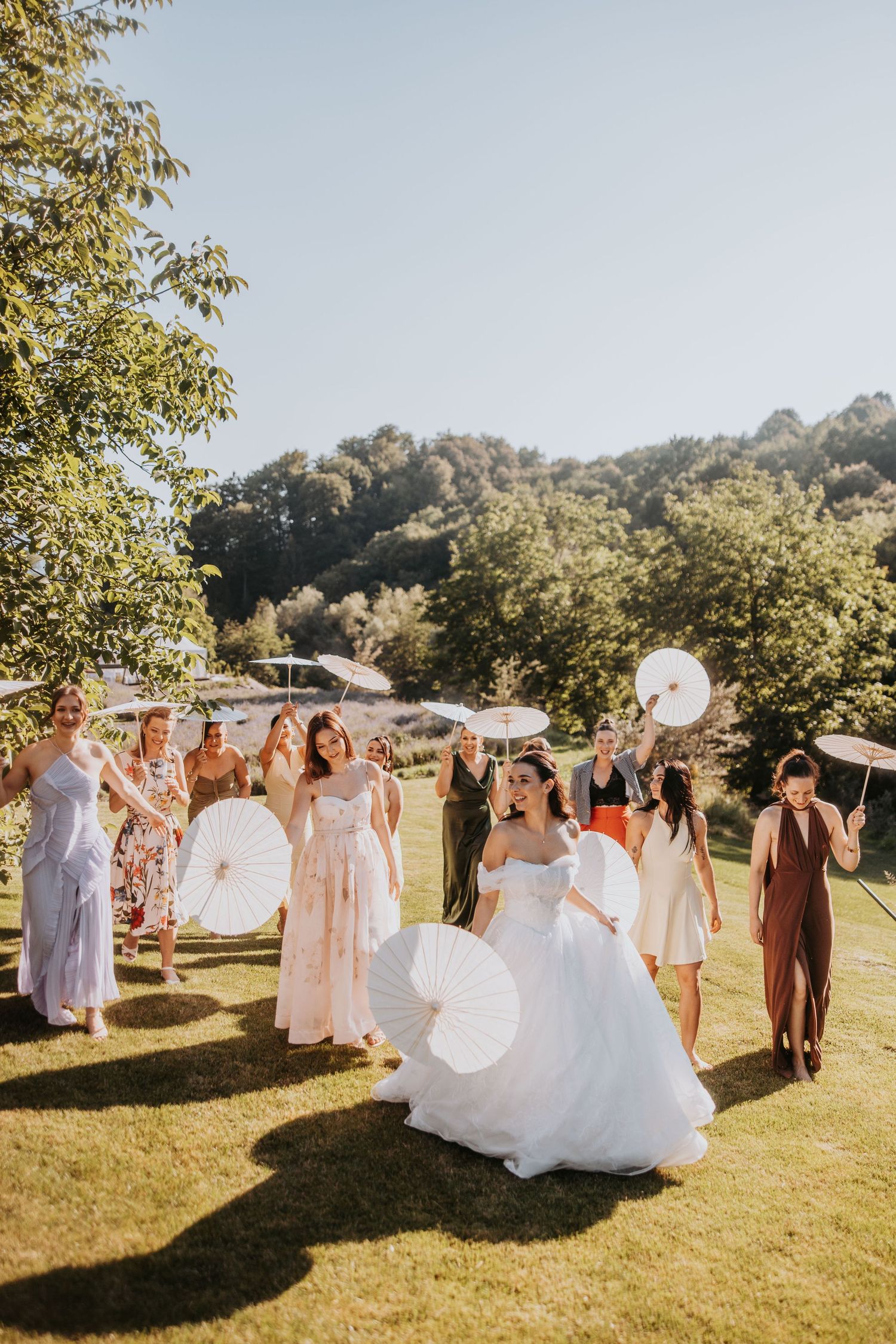 Bride in white gown walks with bridesmaids holding parasols on sunny outdoor wedding day.