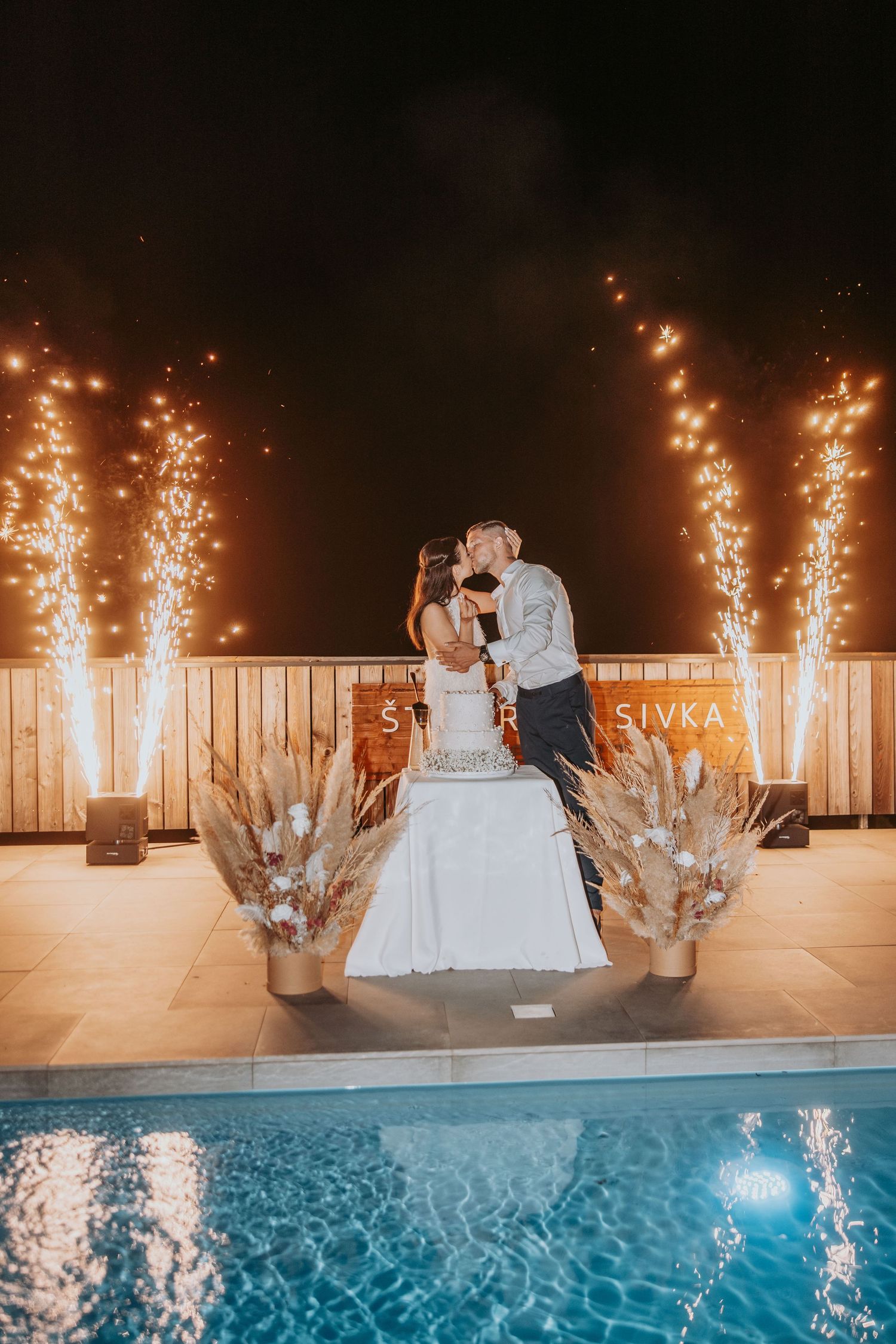Bride and groom kissing by wedding cake with sparkling fireworks beside a pool at night.