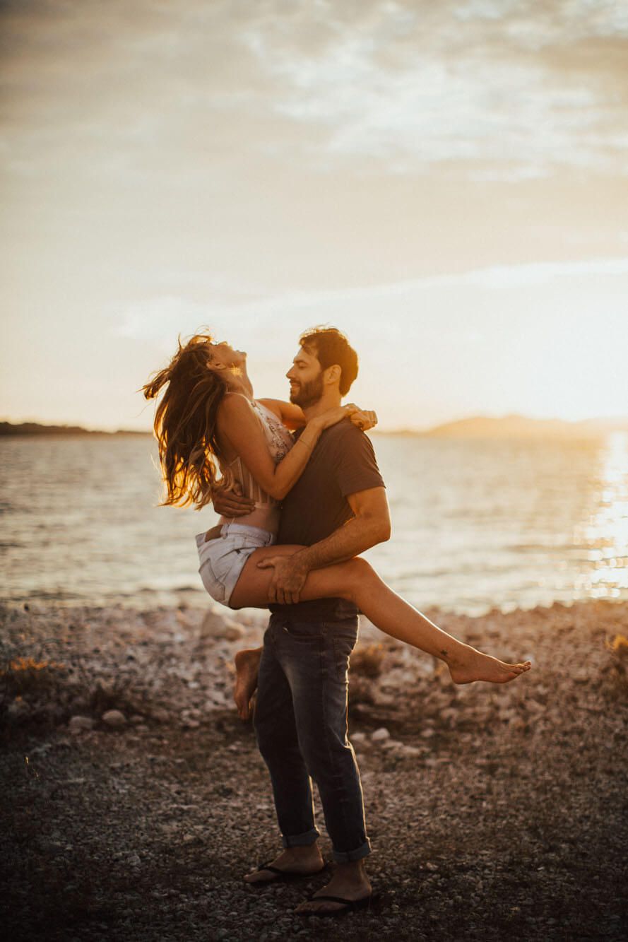 Man lifting woman in his arms on rocky beach at golden sunset by the sea.