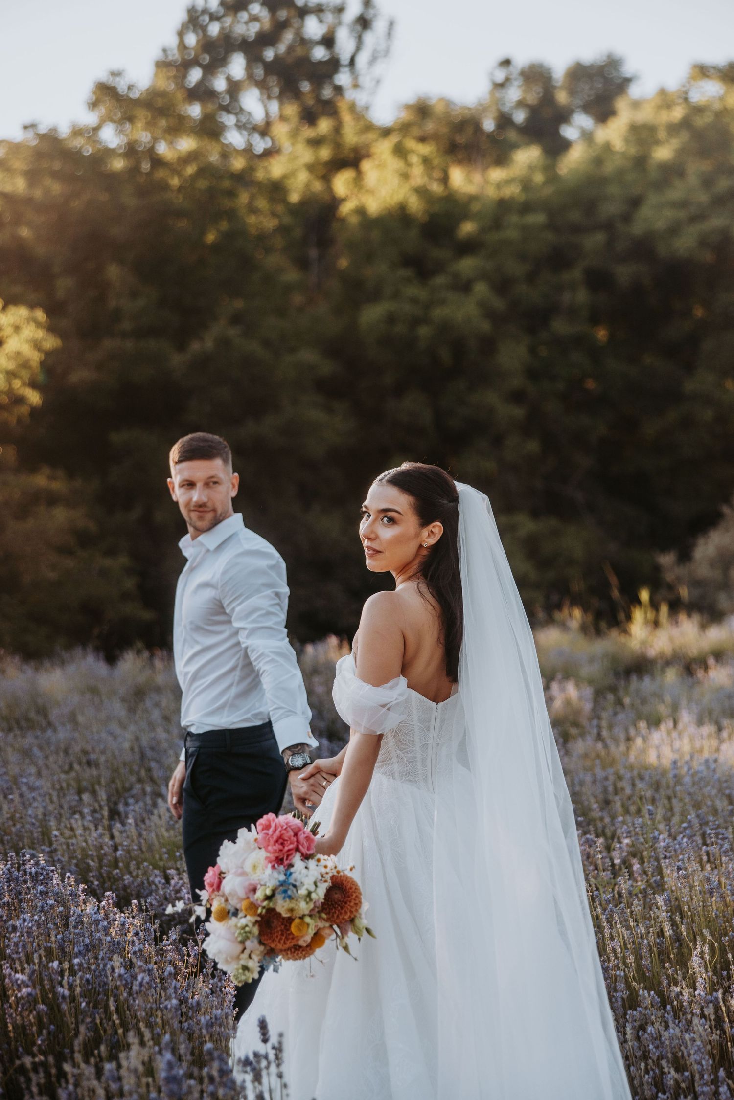 Bride in off-shoulder gown holding colorful bouquet with groom in lavender field at golden hour wedding.