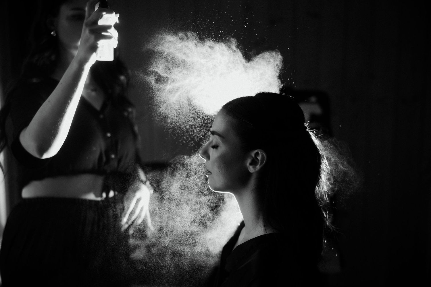 Black and white photo of a makeup artist spraying hairspray on a model backstage, creating a dramatic mist effect.