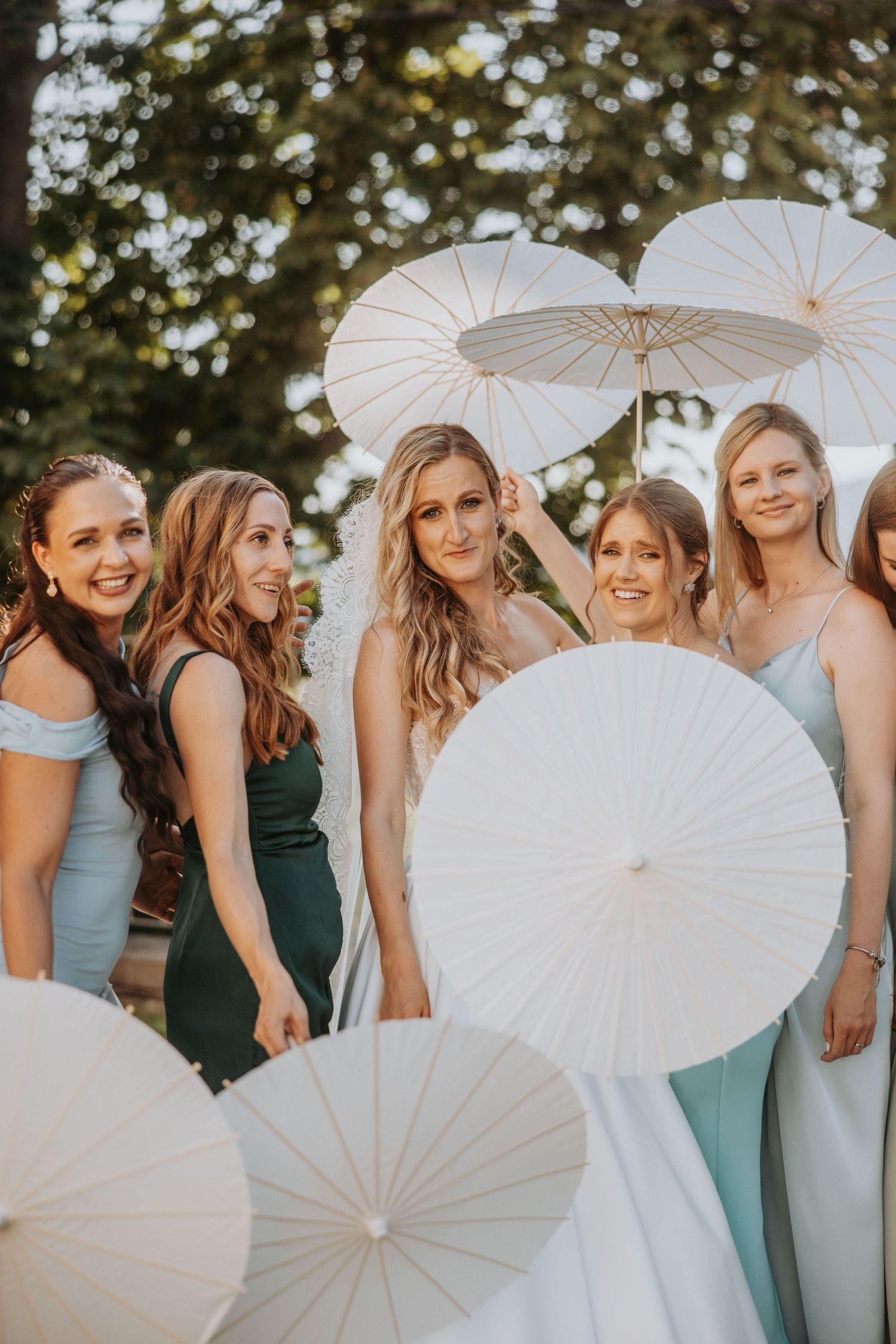 Bride and bridesmaids in mint and green dresses pose with white paper parasols outdoors at wedding.