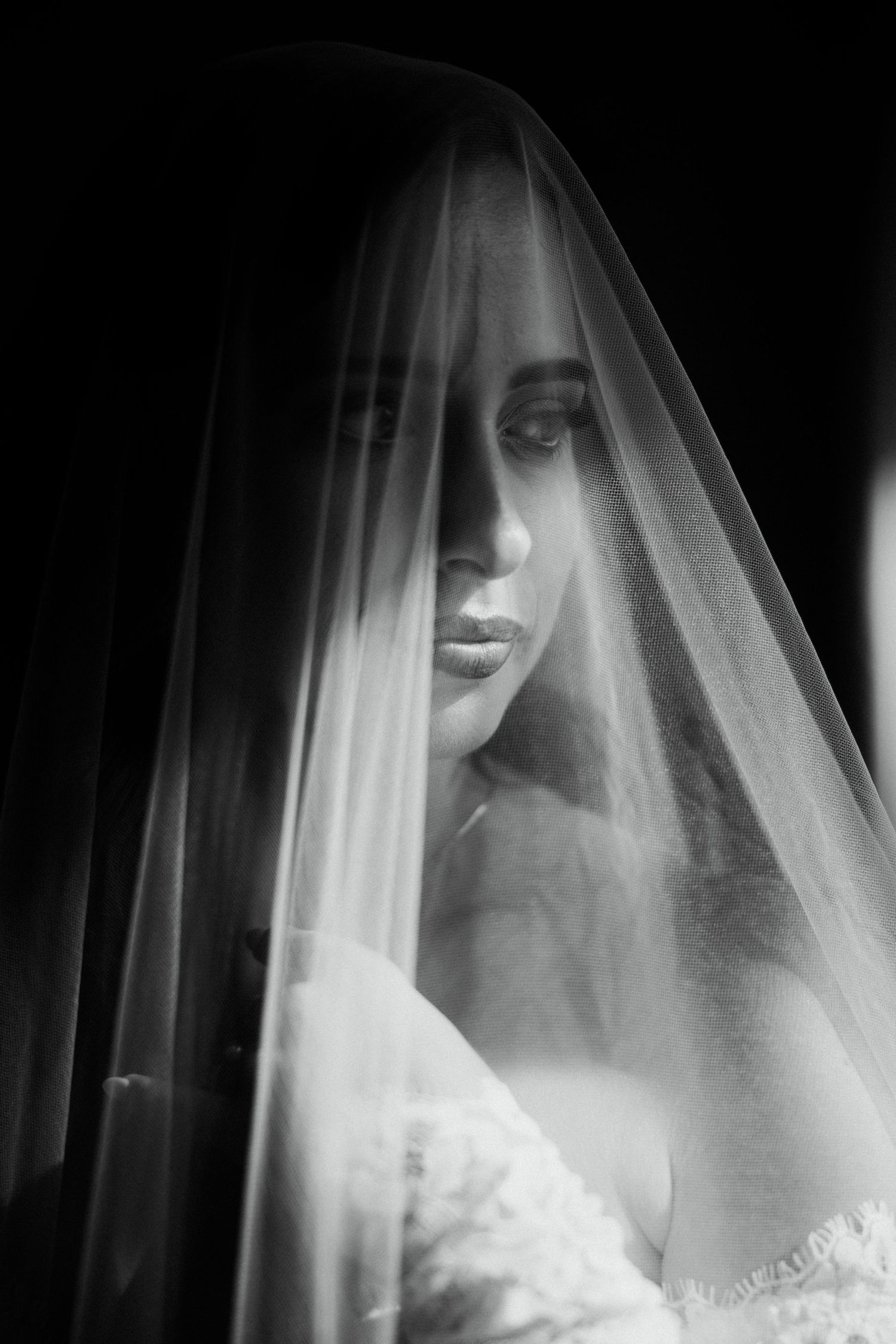 Artistic black and white bridal portrait of bride with veil draped over face in dramatic lighting.
