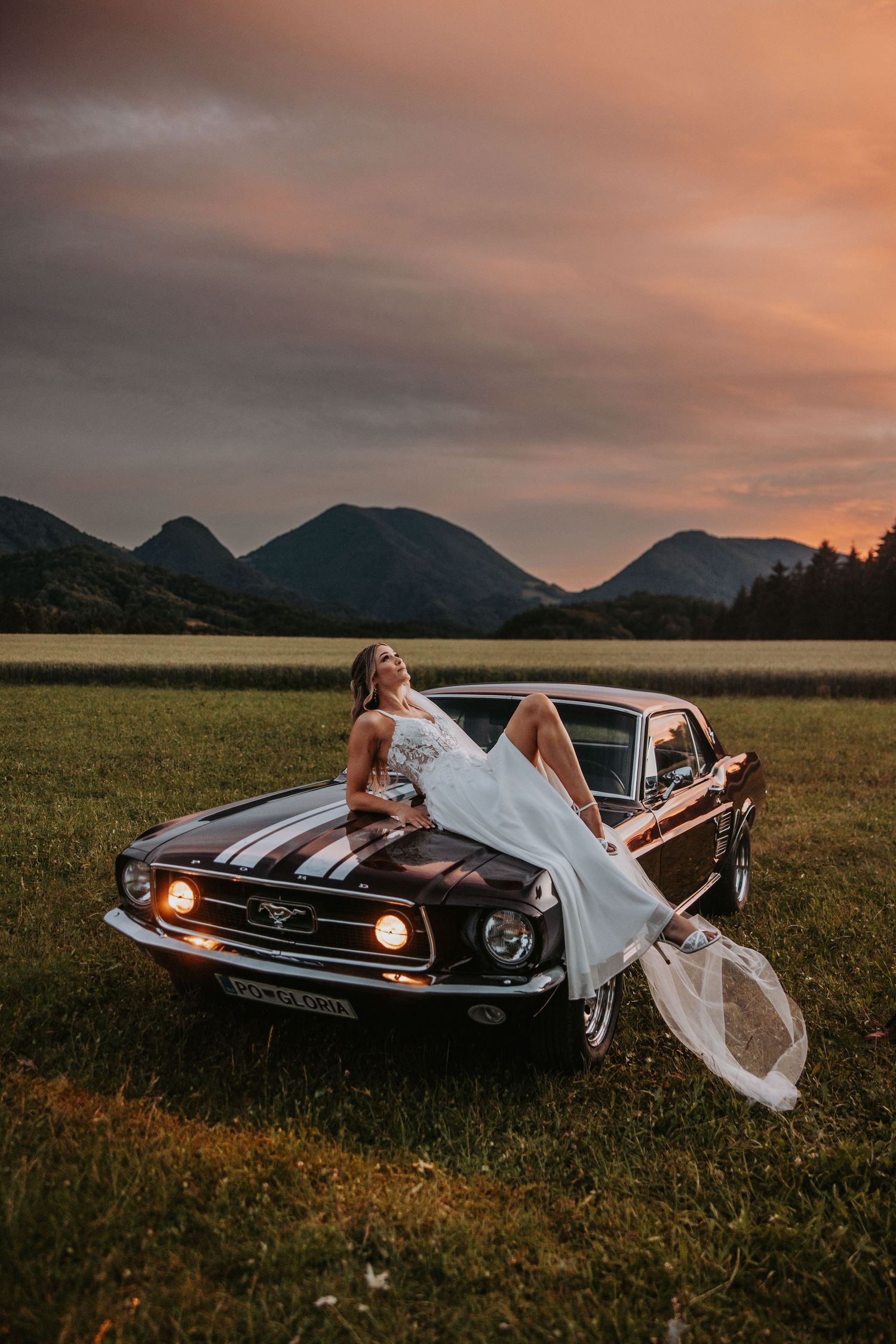 Bride in white gown reclines on hood of vintage Ford Mustang at sunset with mountain backdrop.