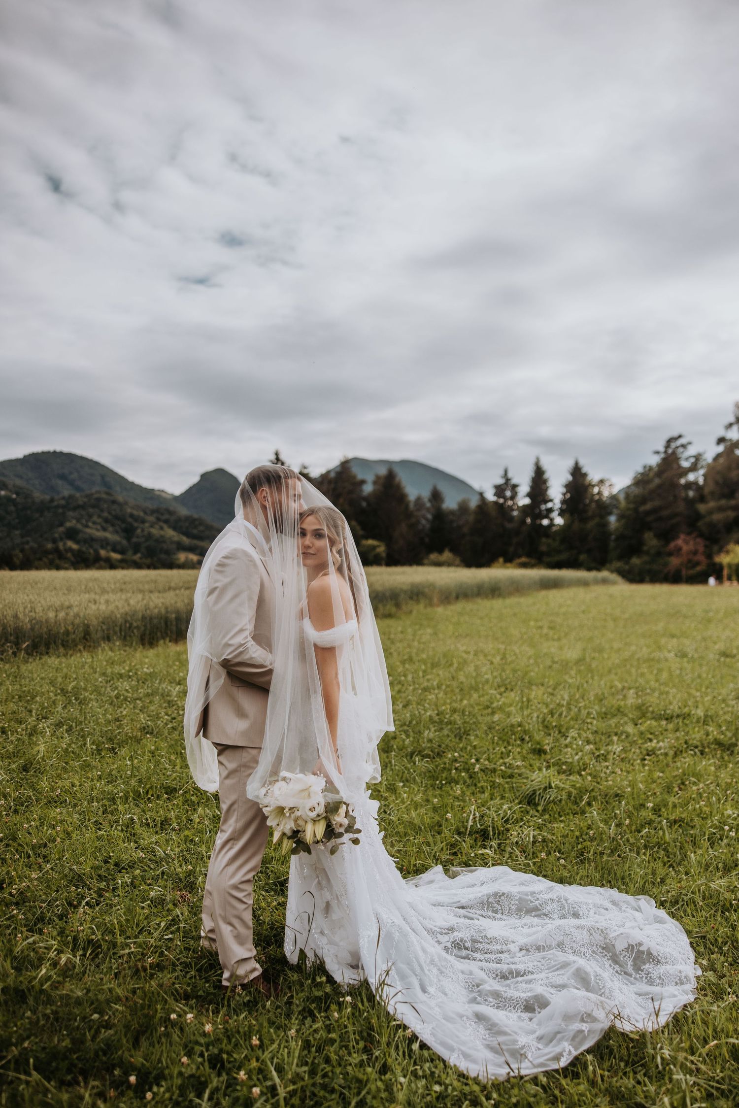 Bride and groom share a kiss under flowing veil in scenic mountain meadow on their wedding day.